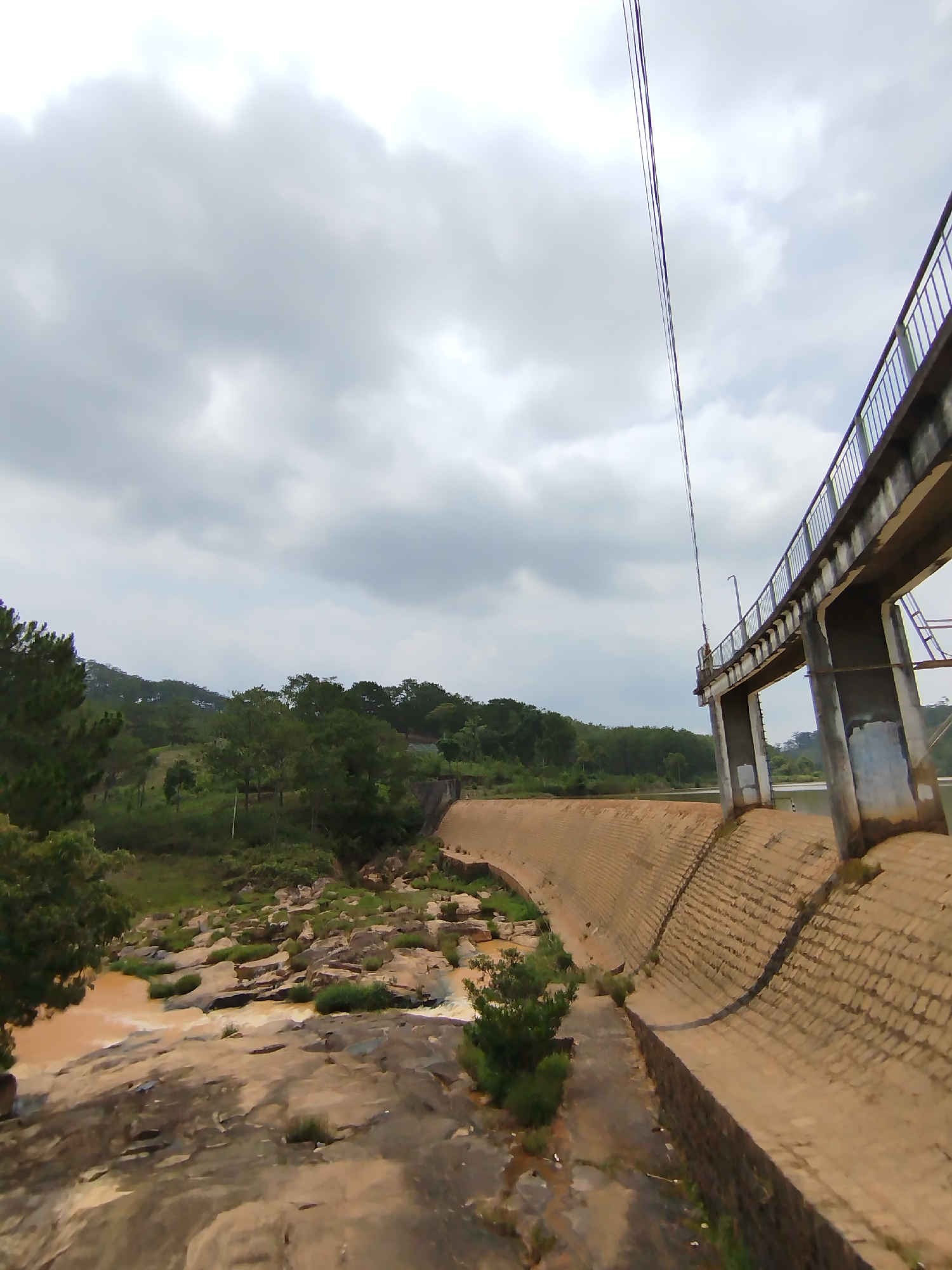 The image presents a wide, slightly low-angle view of a substantial dam structure and its surrounding landscape, identified as being in Lang Biang - Da Lat, Vietnam. The scene is dominated by a curved concrete dam wall, which exhibits a textured, block-like pattern. Above the dam, a concrete bridge or walkway with a metal railing spans across, supported by thick pillars. Utility cables are prominently visible, hanging suspended across the upper part of the frame, seemingly originating from near the bridge.
Below the dam, a river flows with reddish-brown, turbulent water, suggesting sediment content. The riverbed is rocky, with numerous large boulders and smaller rocks partially submerged or exposed. Lush green vegetation, including dense trees and shrubs, covers the hills and banks around the river and dam. On the left side of the frame, a cluster of pine-like trees stands tall, framing the natural elements of the scene.
The sky is overcast with heavy, grey clouds, indicating a cloudy or possibly gloomy day. No people are visible in the image, and there is no specific activity taking place beyond the natural flow of the river. The overall impression is one of a vast, engineered landscape nestled within a natural, verdant environment. No visible text is present in the image.