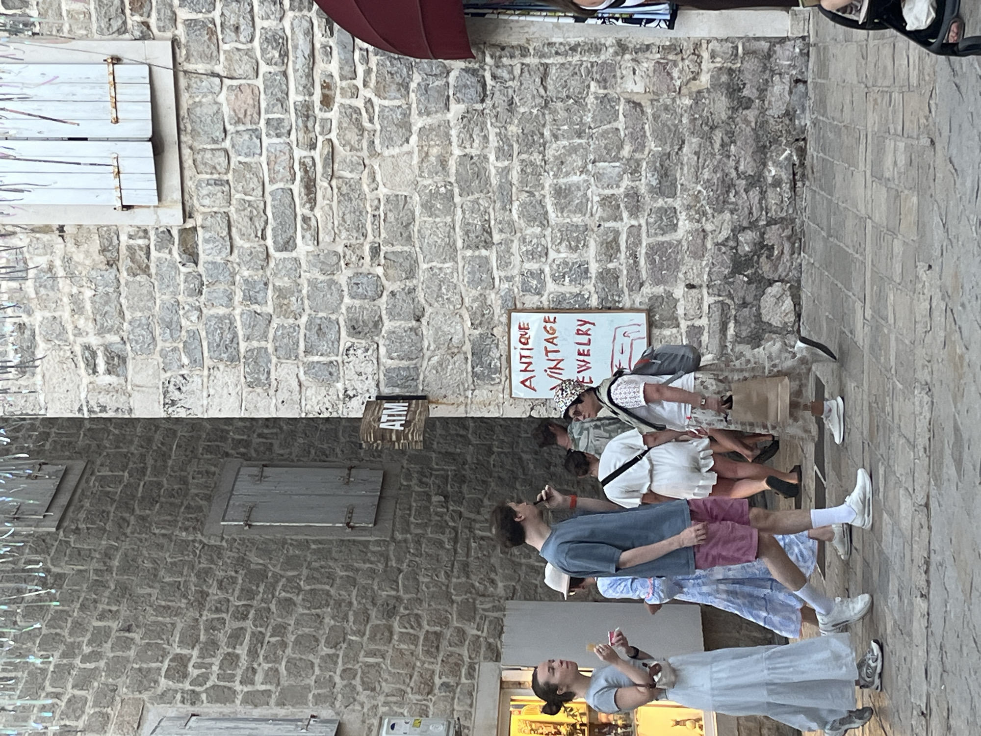 The image depicts a street scene in the Old Town of Budva, Montenegro. Several people are walking on a cobblestone path in front of buildings constructed with rough-hewn stone blocks. One building on the right has a sign that reads "ANTIQUE VINTAGE JEWELRY" in red lettering on a white background, and another sign for an "ATM" is visible on a building in the background. The people are dressed in casual summer clothing, suggesting it is daytime. One woman in the foreground is holding a small container and appears to be eating. Another man is walking past her, looking to his right. The architecture and the cobblestone street are characteristic of the historic areas found in Budva's Old Town.