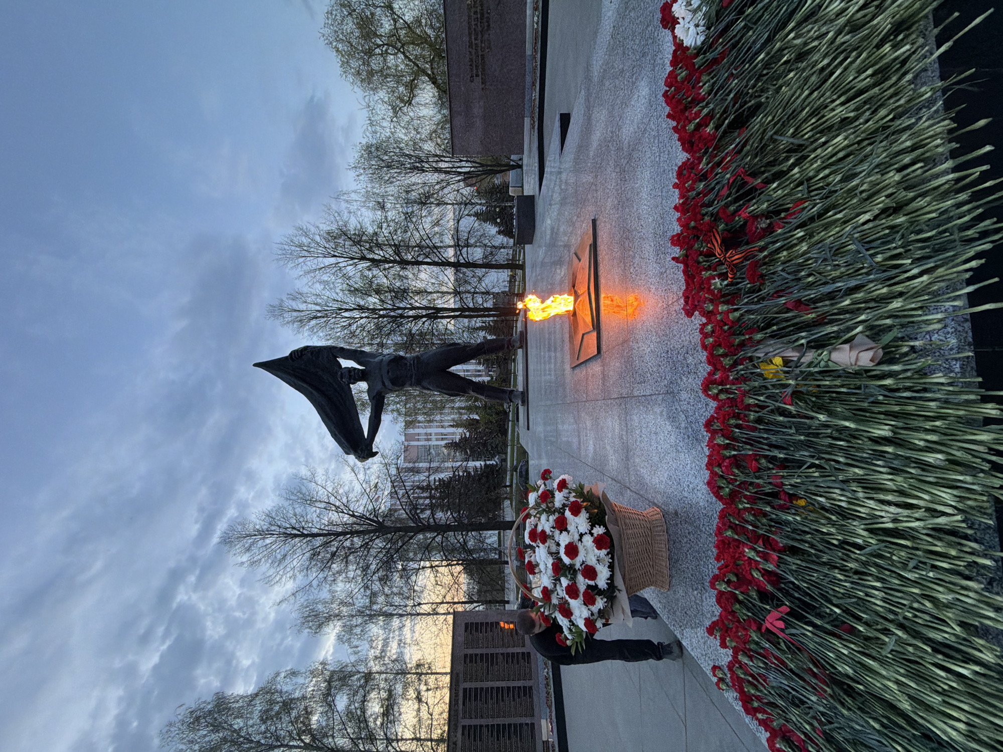 The image depicts a memorial complex in Izhevsk, Russia. A statue of a man holding a flag stands next to an eternal flame burning in a star-shaped fixture. A flower basket and numerous red carnations have been laid at the base of the memorial. A person is partially visible placing the flower basket. The sky is overcast, and bare trees surround the memorial. A building and a wall with inscriptions can be seen in the background.