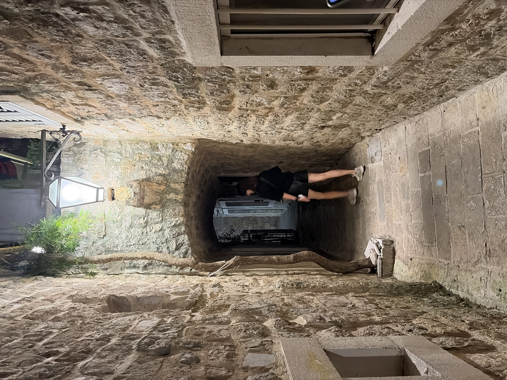 The image depicts a narrow, stone-walled alleyway illuminated by a single overhead lamp. A young woman with dark hair, wearing a black t-shirt and black shorts, is walking away from the camera towards an arched opening at the end of the alley. She is wearing white sneakers. The walls are constructed from rough, uneven stones, typical of historic architecture. A thin, vine-like plant grows up the left wall, reaching towards the lamp. The ground is paved with cobblestones. The setting appears to be the Old Town of Budva, Montenegro, a common characteristic of its ancient streetscapes.