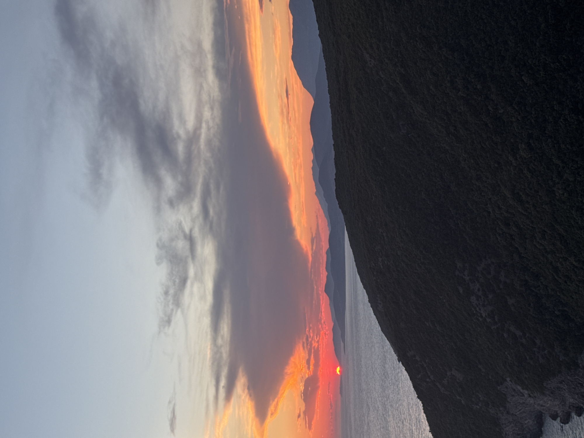 This is a landscape photograph taken at sunset in the city of Zagora, Montenegro. The image shows a view of the Adriatic Sea with mountains in the background. The sun is low on the horizon, casting a vibrant orange and red glow across the sky and clouds. The sea is calm, reflecting the colors of the sunset. In the foreground, a dark, tree-covered hillside slopes down towards the water. There are no people or objects visible in the scene, and no actions are taking place. The overall mood of the image is peaceful and serene.