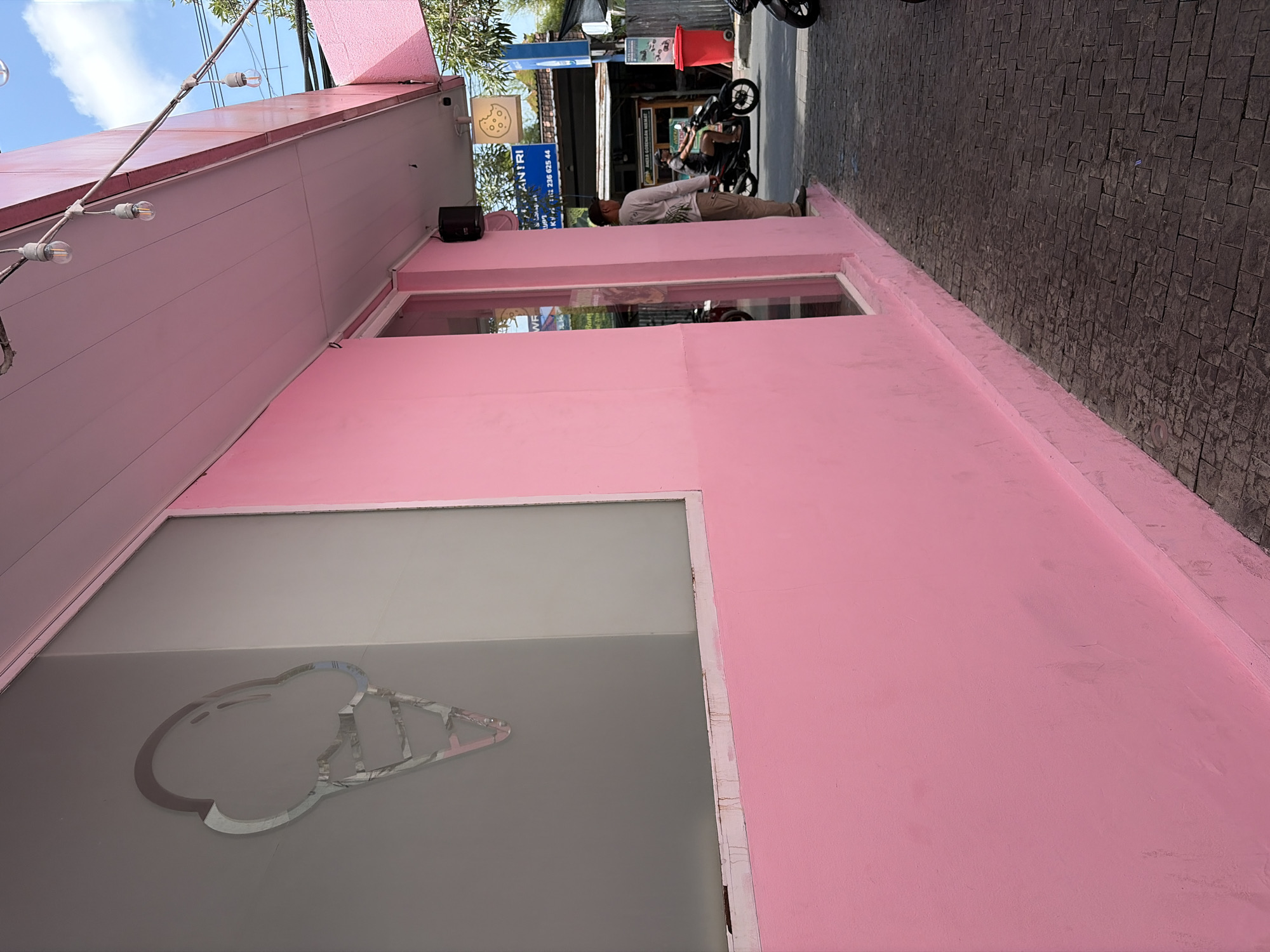 A wide shot captures a vibrant pink building with a large window displaying an ice cream cone decal. The building has a white awning with hanging lights. A person stands near the entrance, looking out at the street. To the right, a paved sidewalk leads to a street where a person rides a motorcycle. The background shows a clear blue sky and some greenery. The scene is located in North Kuta, Indonesia, likely a commercial area given the presence of shops and street traffic.