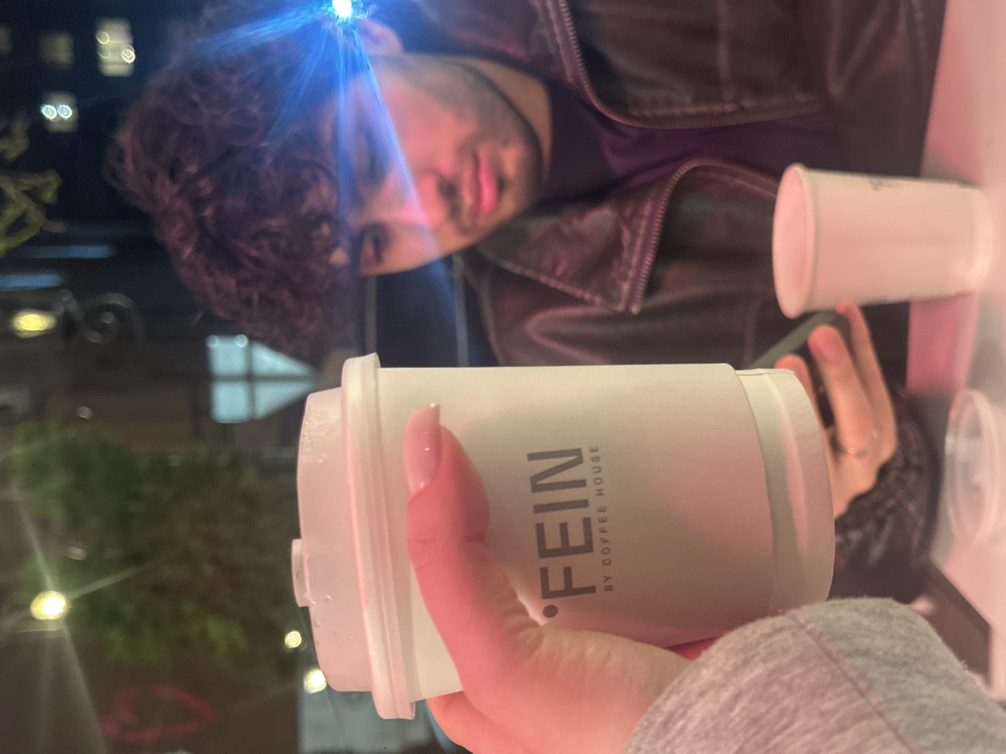 The image depicts a casual indoor scene, likely in a cafe in Yerevan, Armenia, during the evening or night. The perspective is from a person holding a coffee cup in the foreground. This white disposable coffee cup prominently displays the text "FEIN BY COFFEE HOUSE" in dark lettering. The hand holding the cup has manicured fingernails and is wearing a light grey, textured sleeve.
In the background, a man with dark, curly hair and a beard or stubble is visible, wearing a dark leather jacket over a dark shirt. His face is partially blurred but he appears to be looking towards the camera. A strong blue light source, possibly a digital screen or an artificial light, creates a bright lens flare on his forehead and reflects off his hair, casting a blue hue. Another plain white cup rests on a light-colored table in front of him. The background behind the man shows blurred lights, indicating an outdoor urban setting viewed through a window at night. The overall impression is of a relaxed social interaction or a coffee break.