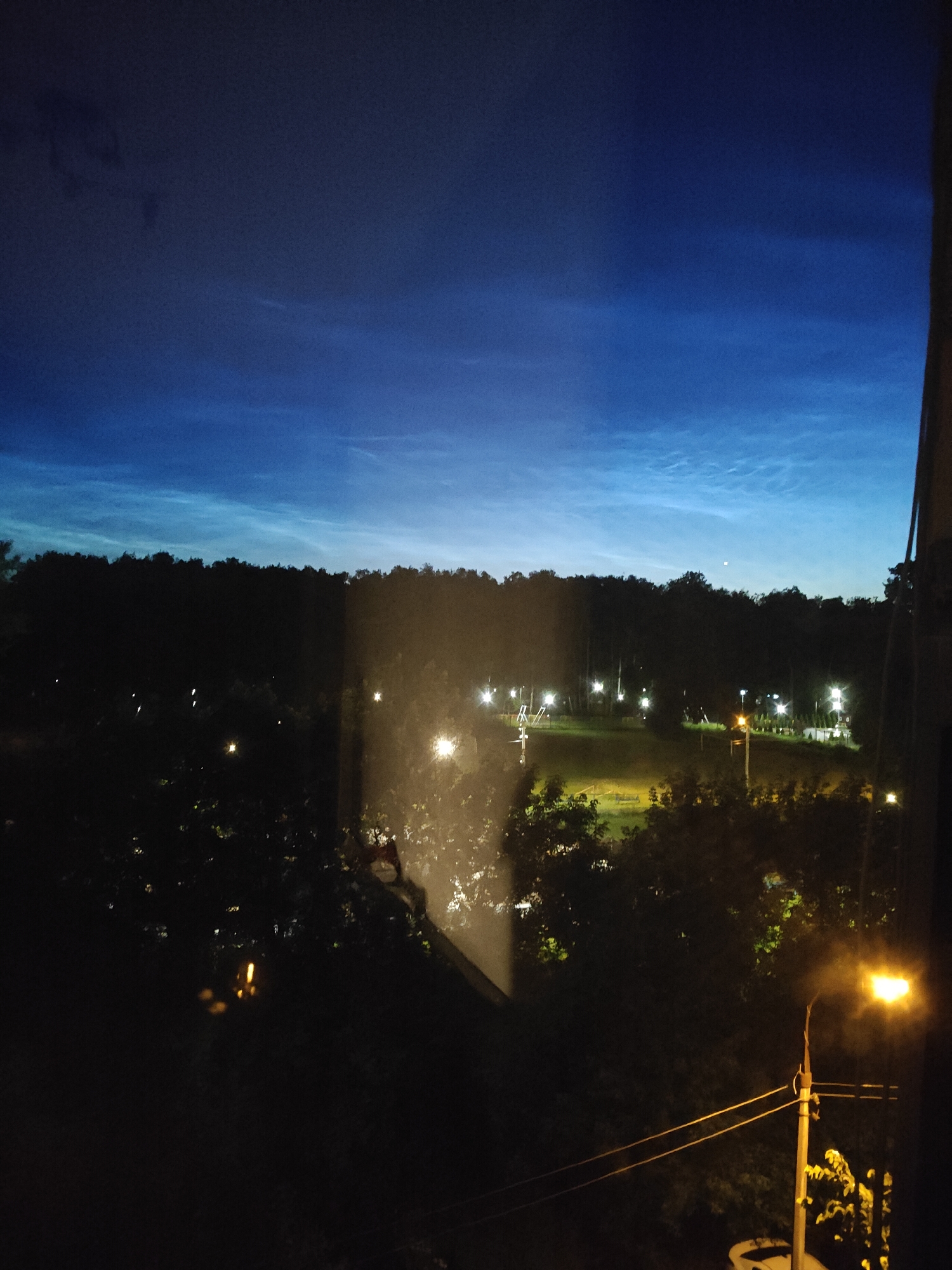 The image captures a nighttime view from a window, overlooking a park-like area with trees and lit lampposts. The sky is a deep blue, transitioning to a lighter shade near the horizon, with wispy clouds visible. In the foreground, the window itself reflects light, obscuring some of the view. Beyond the trees, a park is illuminated by numerous streetlights. Some of these lights highlight playground equipment, suggesting a public recreational space. The overall impression is one of a quiet, urban night scene in Pushkino, Russia.