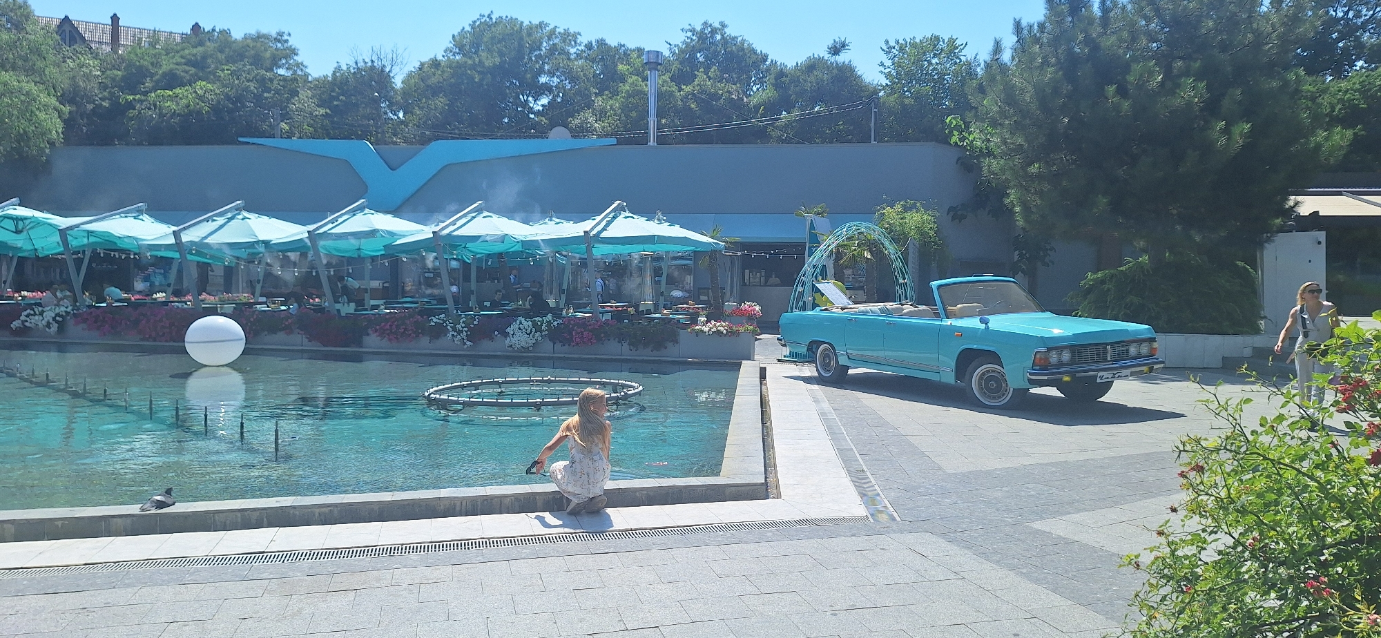 In an outdoor urban setting, a blue convertible car is parked on a paved area. A woman in a light-colored outfit walks away from the car. In the foreground, a fountain with a white sphere and a circular structure is visible. A pigeon is perched on the edge of the fountain. Behind the fountain, an outdoor seating area with turquoise umbrellas and tables is set up, with flowering plants in front. Trees and a building with a blue architectural element are in the background. The sky is clear and blue.
