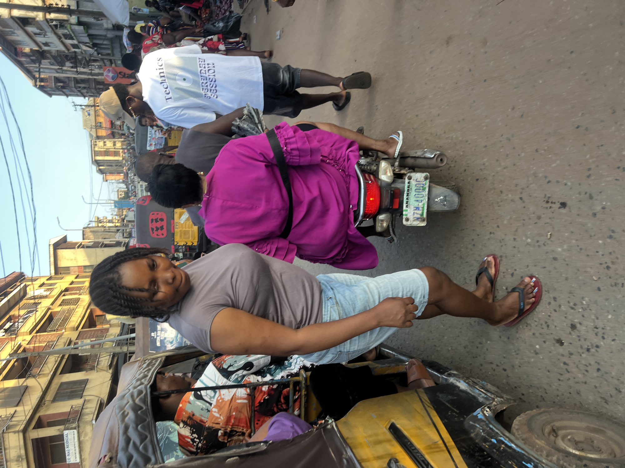 The image captures a street scene in Nkpor, Nigeria, characterized by a busy thoroughfare and bustling pedestrian activity. In the foreground, a woman wearing a grey t-shirt and denim shorts stands on the right side of a yellow auto-rickshaw, her gaze directed downwards. Next to her, a woman in a vibrant fuchsia dress is seated on a motorcycle, her back to the camera, and her feet adorned with flip-flops. The motorcycle is equipped with a license plate that reads "ANAMBRA NZM-4000C."
Behind the woman in the grey t-shirt, another auto-rickshaw is partially visible, its interior containing passengers. In the background, a man wearing a beige cap and a white t-shirt with the word "Technics" emblazoned on it walks away from the camera. He is accompanied by other individuals and the scene is lined with multi-story buildings, some appearing to be shops or residences, with balconies and awnings. The ground is paved with asphalt, showing signs of wear and tear. The lighting suggests it is daytime, possibly late afternoon, given the warm, golden hue of the sunlight illuminating the buildings. The overall atmosphere is one of everyday life and commerce in a Nigerian urban setting.