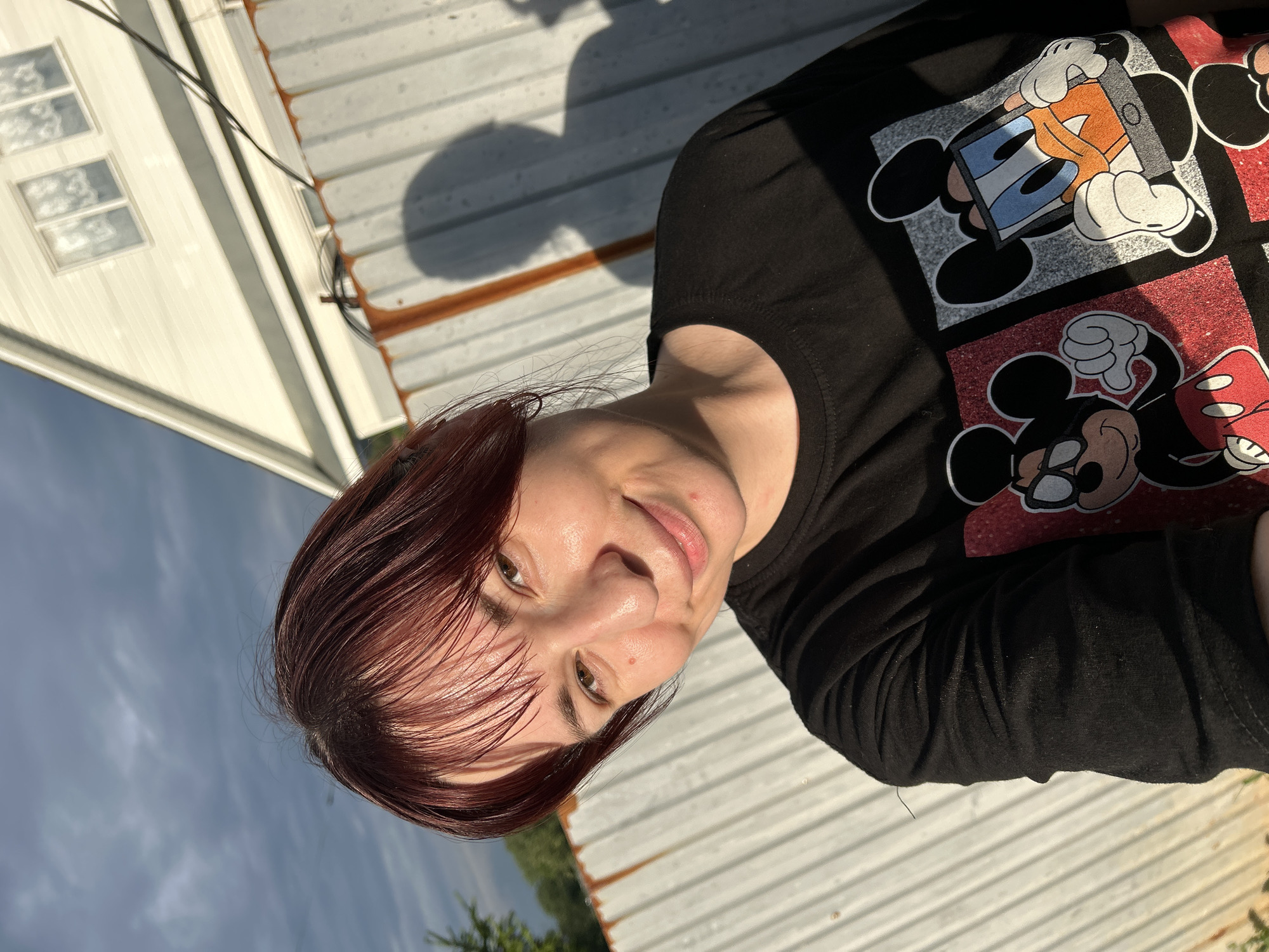 A woman with reddish-brown hair and bangs is taking a selfie outdoors. She is wearing a black t-shirt with a graphic print of Mickey Mouse and Donald Duck. She is standing in front of a corrugated metal fence, with a white house and a cloudy sky in the background. The lighting suggests it is late afternoon or early evening, with the sun casting a shadow of the woman on the fence. The environment appears to be a residential area, possibly in the city of Fokino, Russia, given the architectural style of the house and the presence of utility wires. There are no other individuals or animals visible in the frame.