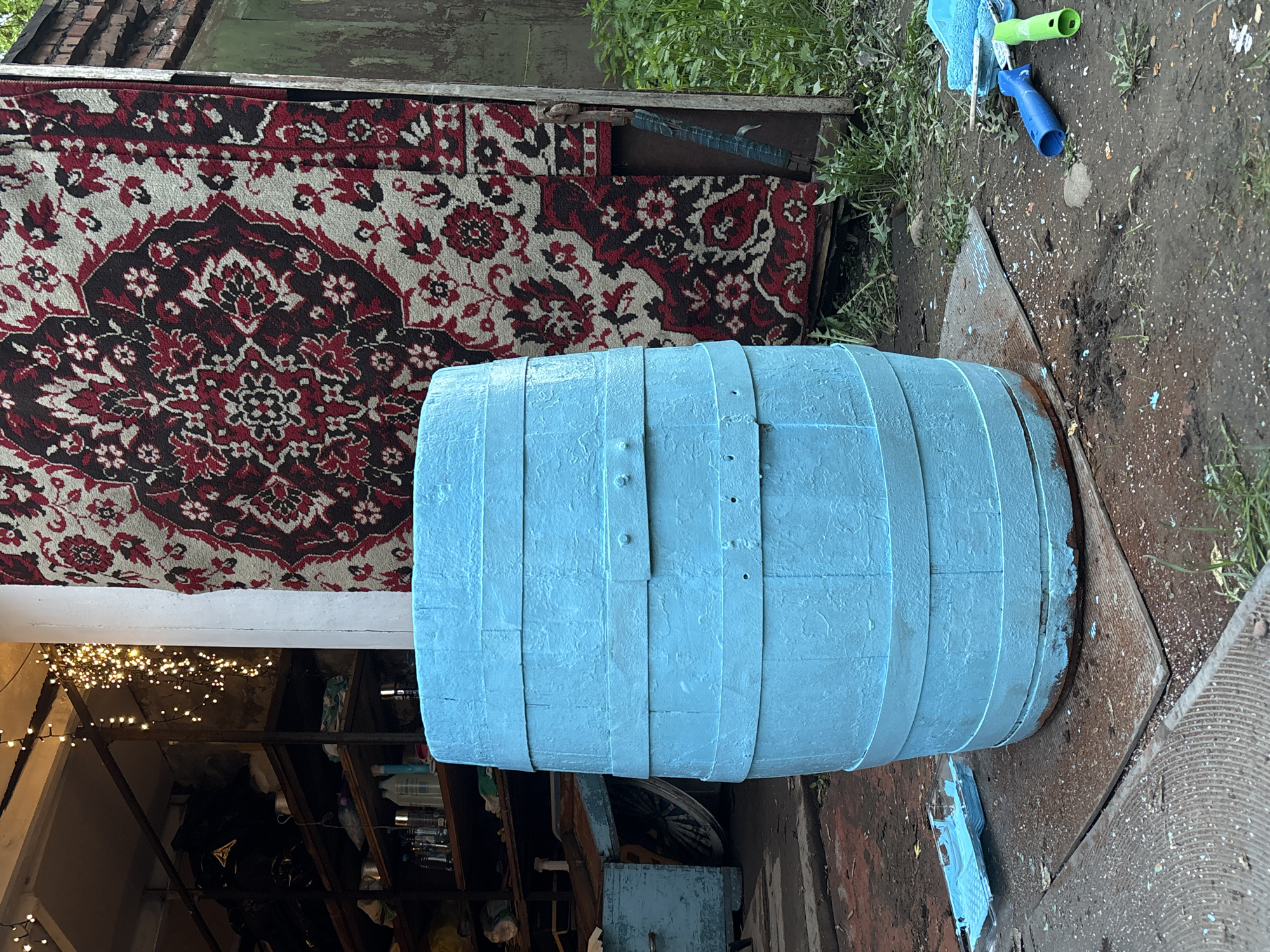 The image shows a blue-painted wooden barrel positioned outdoors on a gray surface. A red and black patterned rug hangs vertically behind the barrel. To the right of the barrel, on the ground, are painting tools: a blue paint roller and a blue cleaning brush. Behind the rug and barrel is a green building. To the left and below the barrel, a shelf is visible, partially obscured, with various items stored on it. String lights are hanging in the area to the left. The location is likely an outdoor area, possibly a yard or storage area, in Krasnoyarsk, Russia.