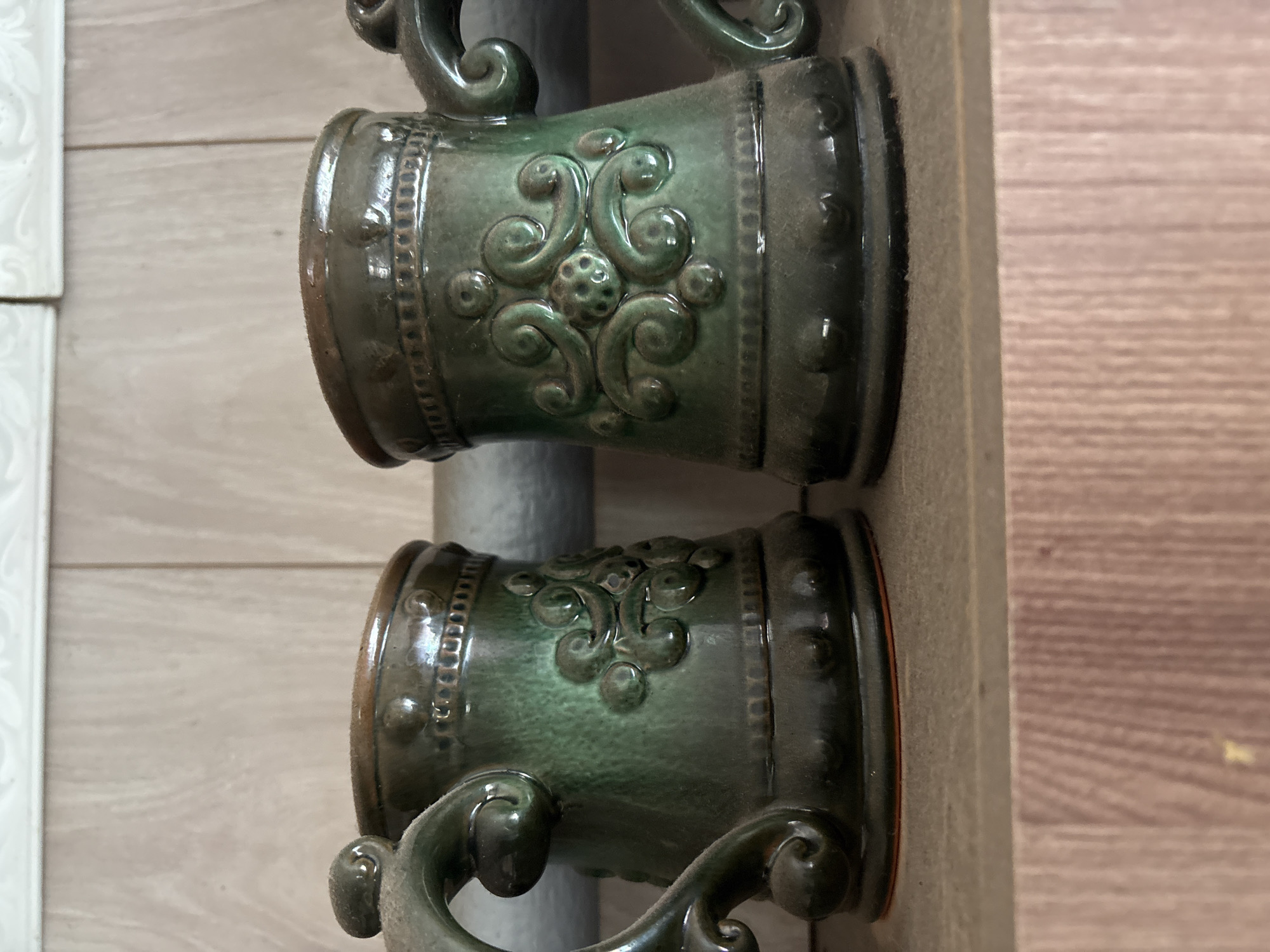 Two green ceramic mugs with ornate carvings and handles are displayed on a shelf. The mugs have a glossy finish and appear to be dusty. The shelf is made of wood, and behind it, there is a textured wall. The overall setting suggests an indoor display or storage area.