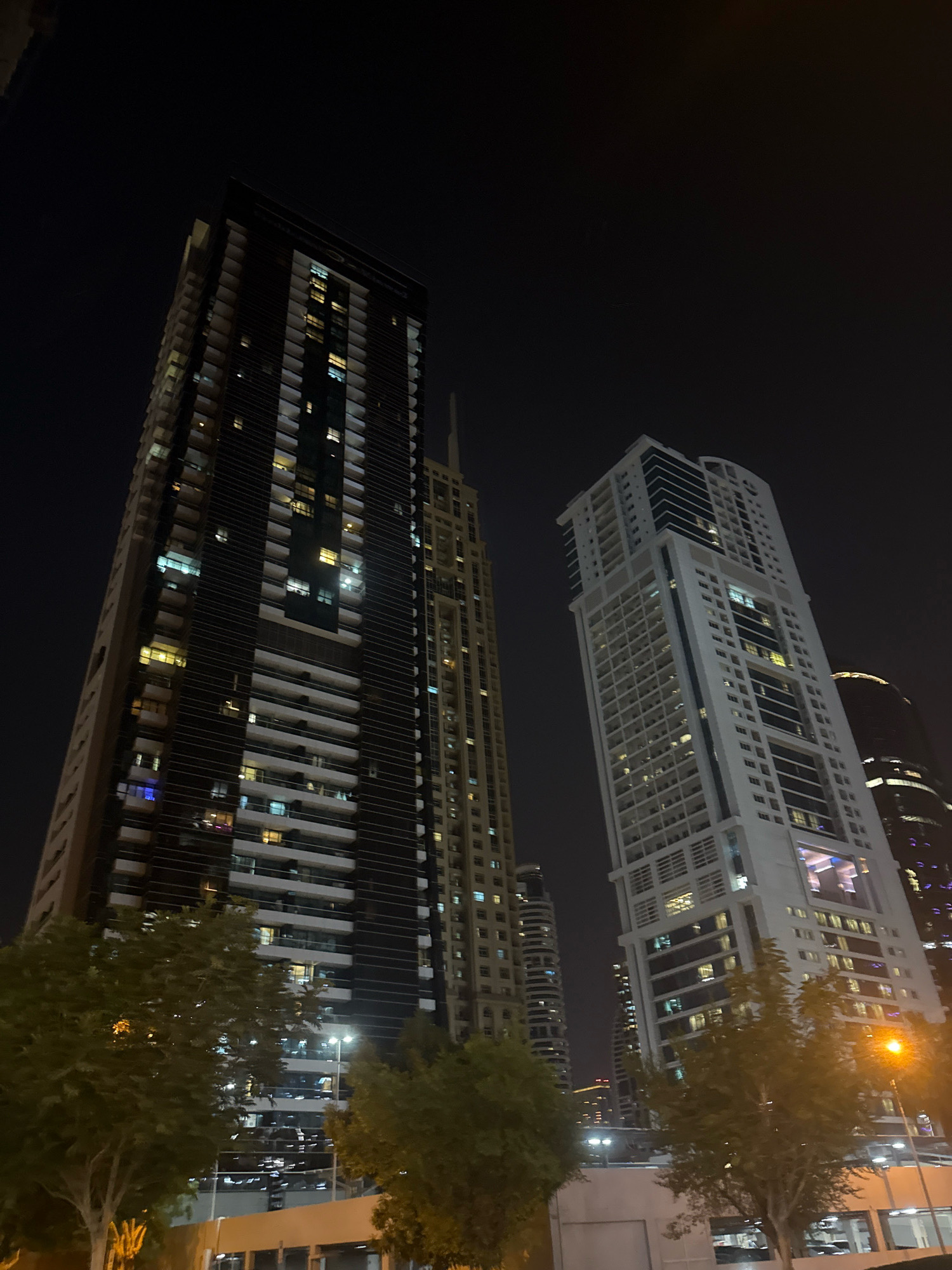 The image captures a night view of modern skyscrapers in Dubai, United Arab Emirates. In the foreground, lush green trees are visible, with a street light illuminated on the right side of the frame. Behind the trees, multiple tall buildings dominate the scene. The building on the left is a dark-colored skyscraper with many floors featuring balconies and lit windows, indicating occupation. To its right, a lighter-colored, slender skyscraper stands tall, also adorned with numerous windows. Further in the background, other high-rise buildings are partially visible against the dark night sky. The architecture is characteristic of Dubai's contemporary urban landscape, emphasizing verticality and modern design.