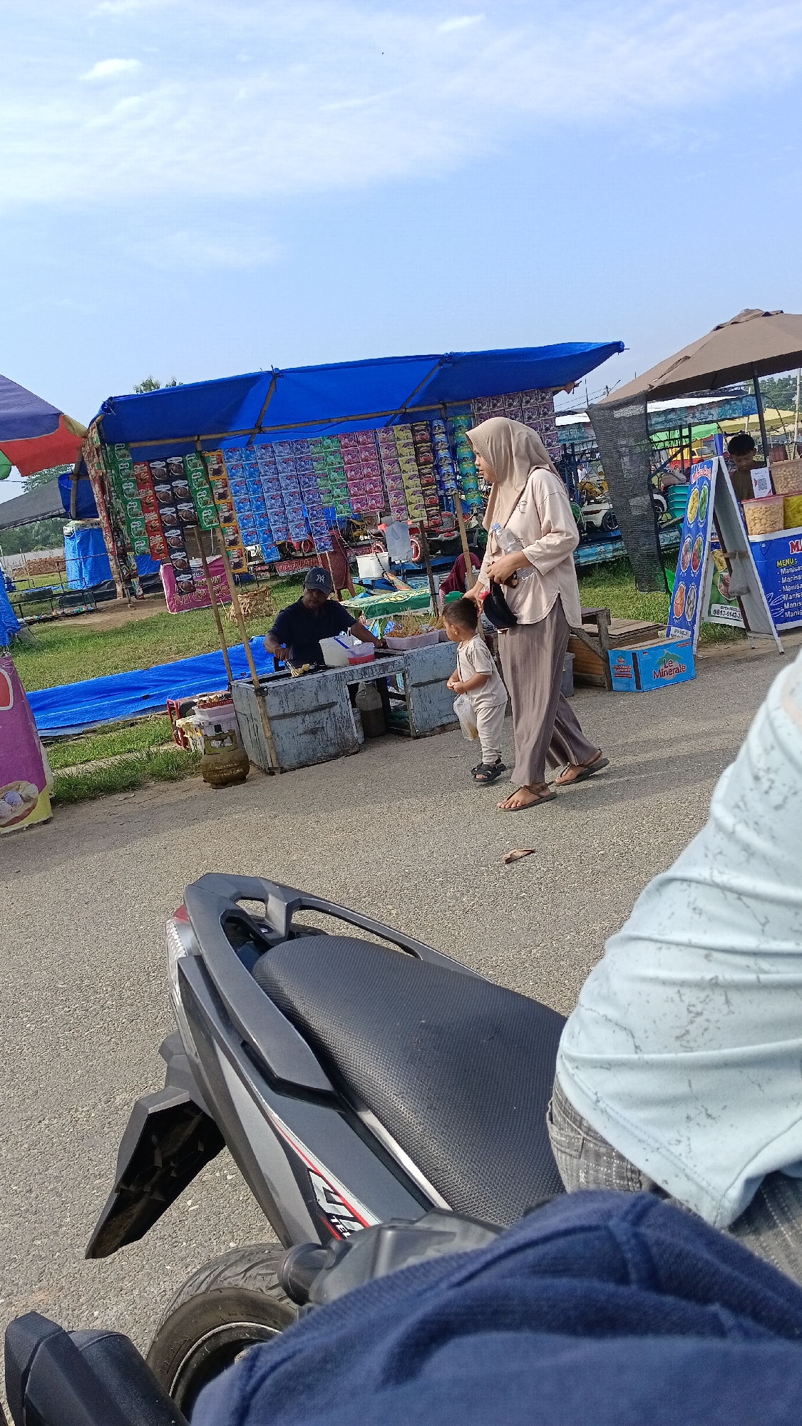 A street vendor is preparing food at a stall, with a woman and a child standing nearby in Aek Kanopan, Indonesia. The vendor, wearing a dark blue cap and shirt, is attending to a grill, while the woman, in a light brown hijab and matching pants, holds a black bag and appears to be watching the child. The child, in a light-colored outfit, stands between the woman and the vendor's stall. The stall is covered by a blue tarp and displays various packaged goods. In the foreground, a portion of a dark-colored motorcycle is visible, suggesting the photo was taken from a rider's perspective. The scene takes place outdoors on a paved surface, with a grassy area and additional stalls visible in the background under a bright sky.