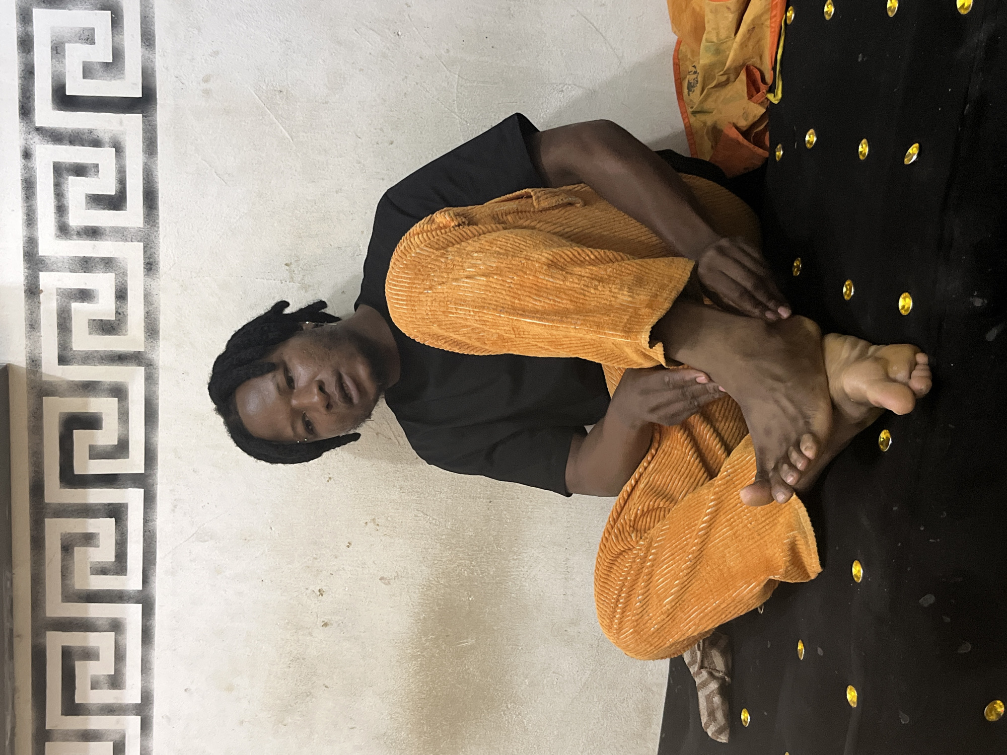 In an indoor setting in Jos, Nigeria, a single adult male with dark skin and dark dreadlocks is centrally positioned, seated on a dark fabric surface. He wears a plain black short-sleeved t-shirt and orange corduroy trousers. He is barefoot, with his left foot crossed over his right, and his hands resting on his lower legs. A small silver-colored stud is visible above his left eyebrow, and a silver-colored earring is present in his left ear. The background consists of a light-colored, possibly off-white, wall exhibiting signs of wear and minor staining. A repeating dark grey geometric meander pattern is stenciled horizontally across the upper section of this wall. The dark fabric surface on which he sits is adorned with numerous scattered, small, oval-shaped, golden, reflective embellishments. To the right of the individual, a folded piece of orange and yellow fabric is visible on the seating surface.