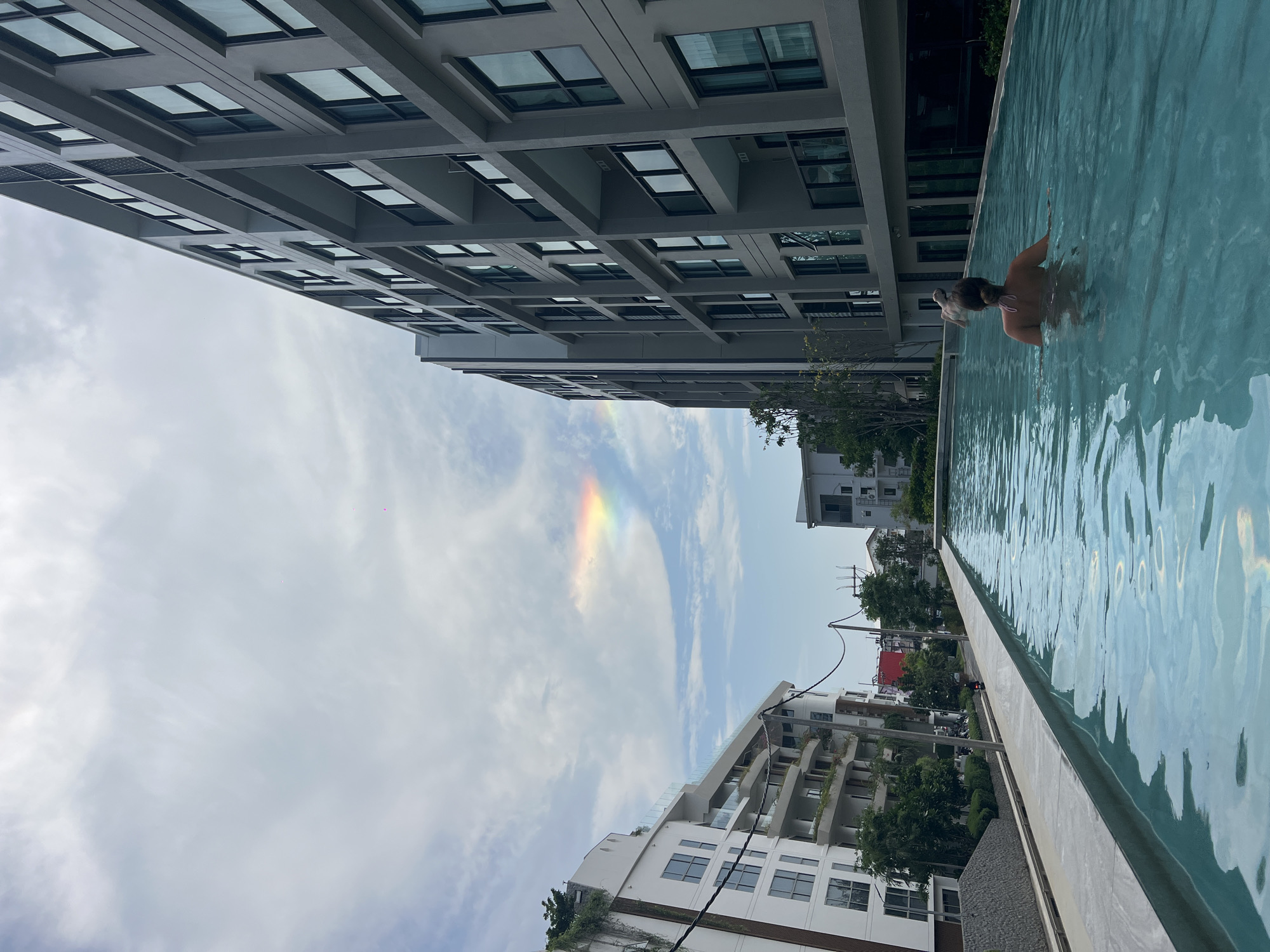 A person is swimming in a pool next to a tall building in Choeng Thale, Thailand. The sky is cloudy with a hint of rainbow. Other buildings are visible in the background.