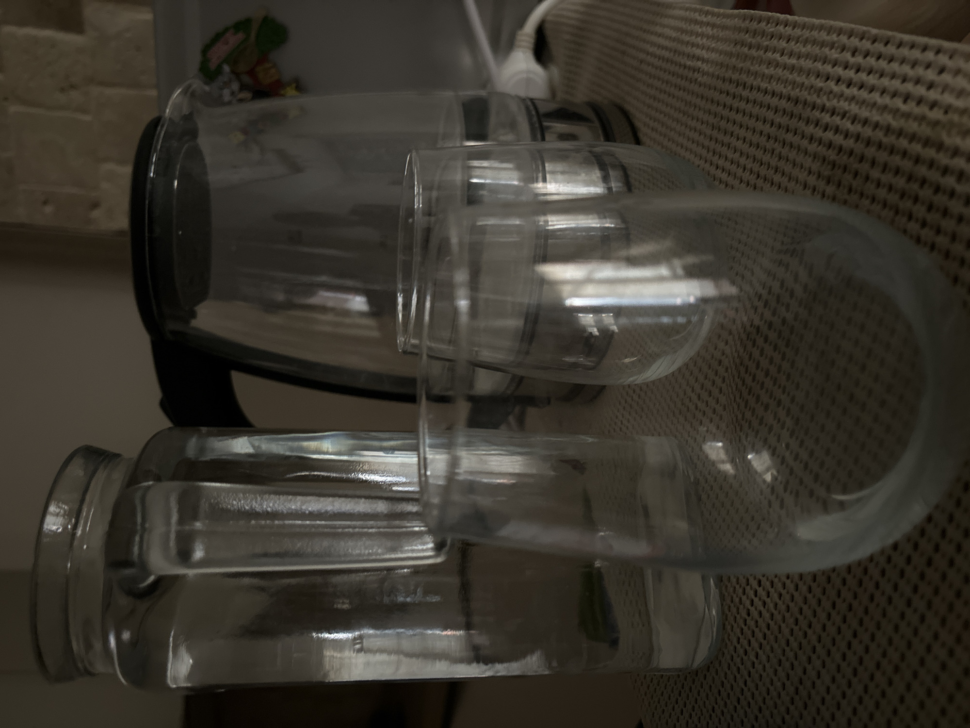 This is a close-up, dimly lit, eye-level shot of glassware and a kettle on a woven placemat. In the foreground, two clear, stemless wine glasses are positioned. The left glass is taller and partially filled with water, showing a slight refraction of light. The right glass is shorter and empty. Behind these, a large, clear glass pitcher filled with water is visible on the left. To the right of the pitcher, a clear electric kettle with a black lid contains water. The kettle is plugged into a white cord that extends out of frame to the right. The background is blurred, but suggests a kitchen environment with tiled wall coverings. The overall impression is of a domestic setting, possibly a kitchen counter or dining table. There are no people present. The scene is located in Budva, Montenegro.
