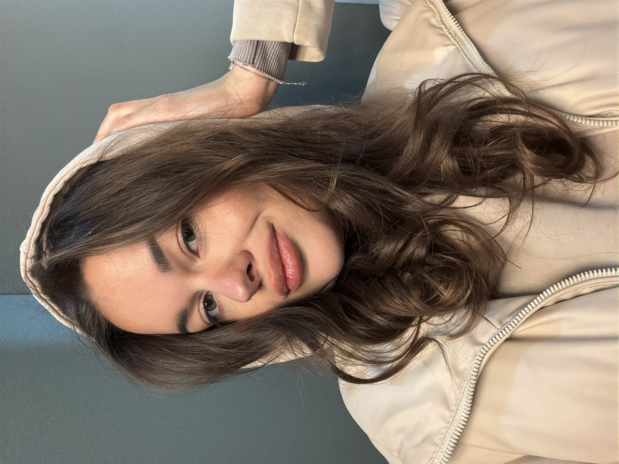 A young woman with brown hair and eyes is positioned in front of a gray wall. She wears a light brown hooded jacket. Her left arm and hand are raised, holding the hood of the jacket. A bracelet adorns her left wrist. The scene's location is not discernible as Krasnoyarsk, Russia.