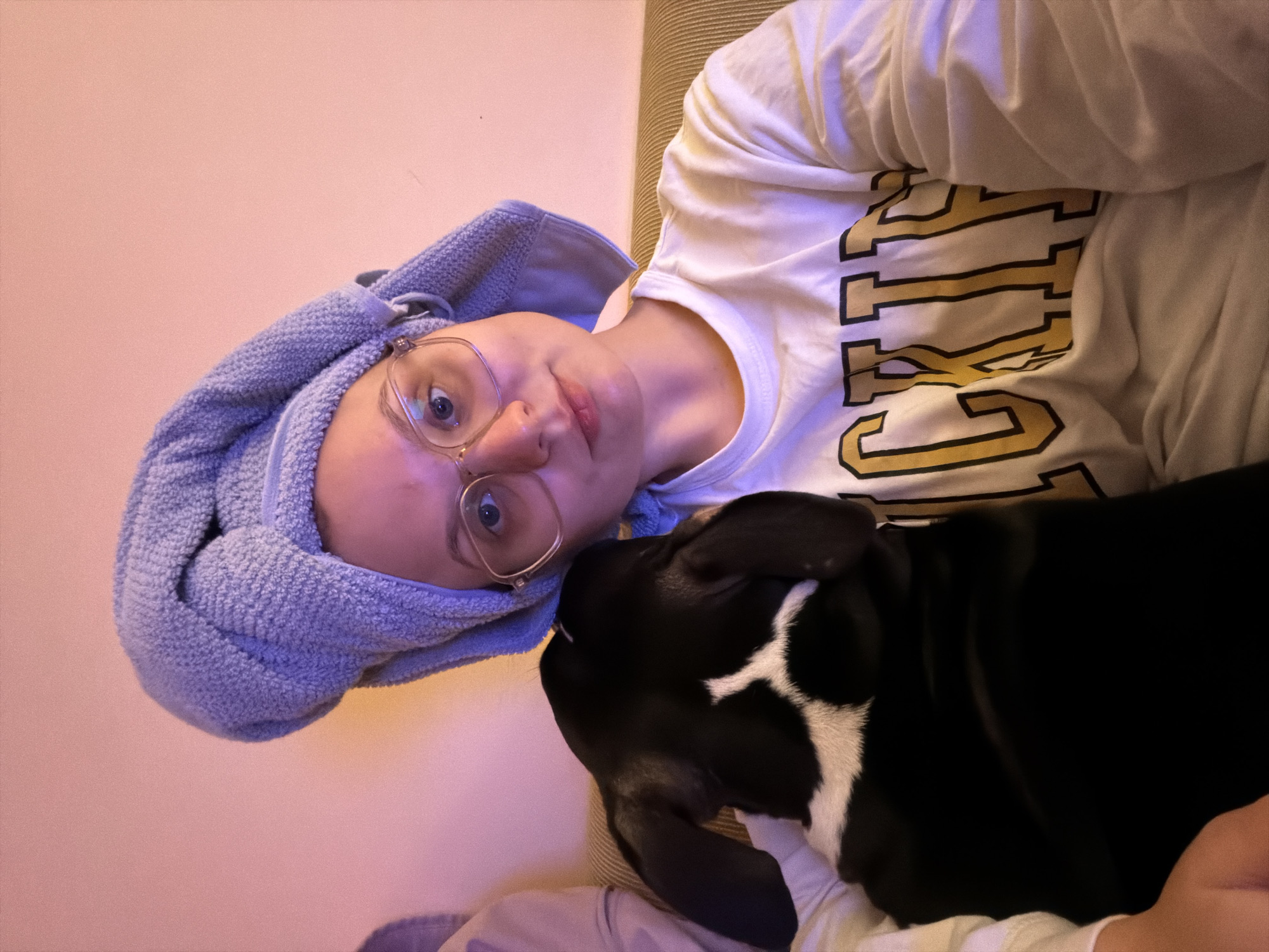 A photograph shows a person and a dog. The person is wearing a light-colored shirt with yellow and black letters on it and has a light blue towel wrapped around their head. They are also wearing glasses. The dog is black with white markings on its chest. The photograph appears to have been taken indoors. It is not possible to determine the location of the photograph as Tel-Aviv, Israel, or any other city based on the information provided.