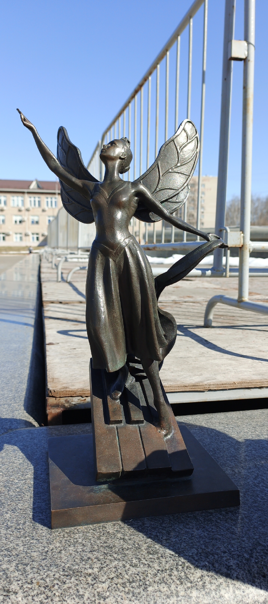 A full-shot image depicts a bronze statue of a female figure with wings, presumably an angel or fairy, set against an outdoor urban backdrop. The statue stands atop a base with horizontal bars, potentially representing piano keys. The figure has both arms outstretched in a gesture of liberation or dance.
The backdrop features a wooden deck with a metal railing. Beyond the railing, a building with rows of windows is visible, suggesting an institutional or residential structure. The sky is clear and blue, indicating a sunny day. Patches of snow can be seen in the background.
The location is the city of Chaikovsky, Russia.
