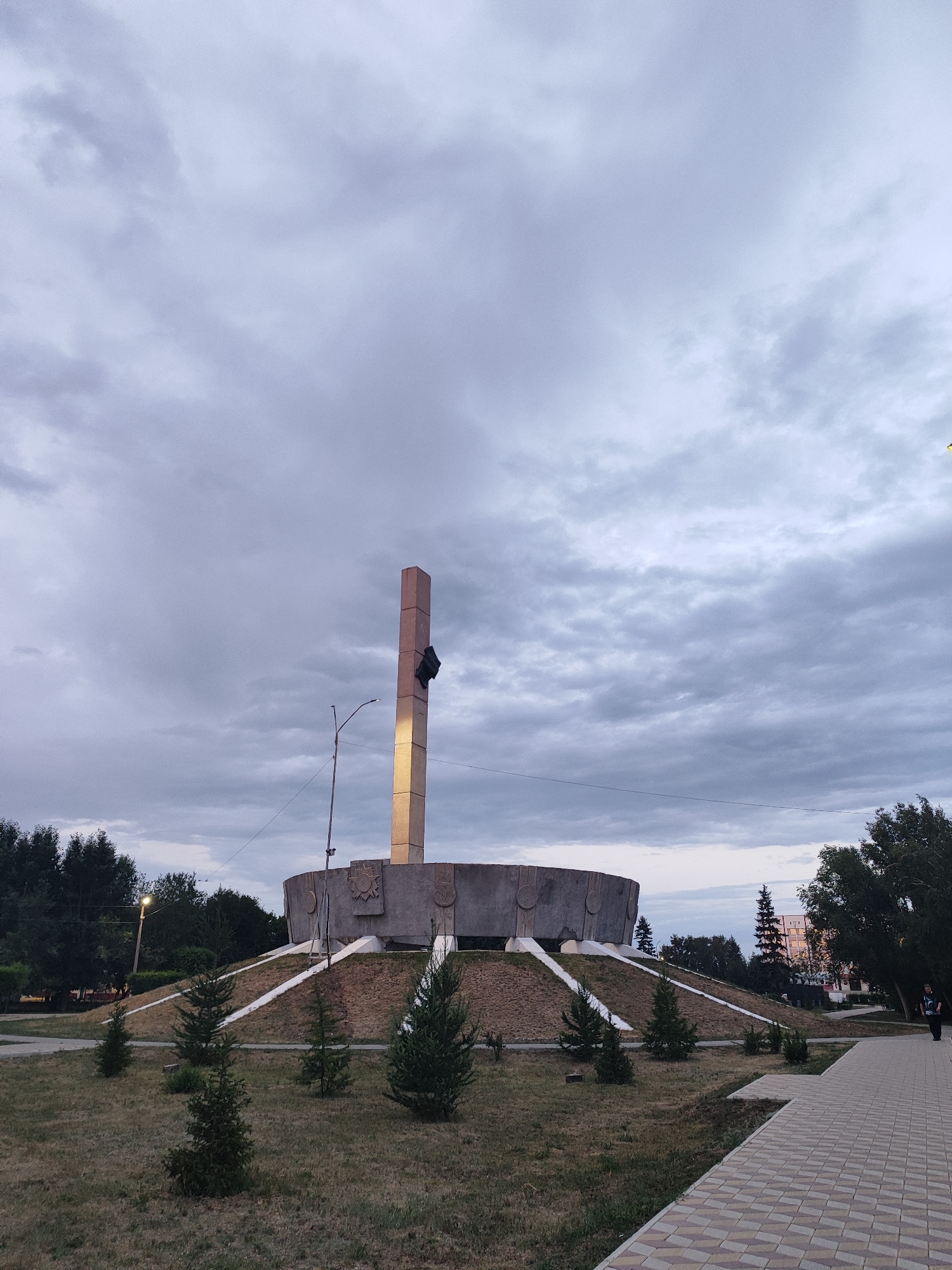 This image captures an outdoor scene featuring a large, concrete monument. The monument is characterized by a wide, circular base elevated on a grassy mound, with a tall, rectangular pillar rising from its center. The pillar is illuminated by a warm light, highlighting a dark object attached to its side. The monument is surrounded by a paved walkway and sparse evergreen trees. In the background, a line of trees and some buildings are visible. The sky is overcast with dramatic clouds, suggesting either early morning or late evening. The overall mood is somewhat somber due to the cloudy weather and muted colors, but the illuminated monument adds a focal point of interest. A single person is visible on the far right of the image, walking away from the monument. The setting appears to be a public space, possibly a park or memorial area. There is no visible text in the image.