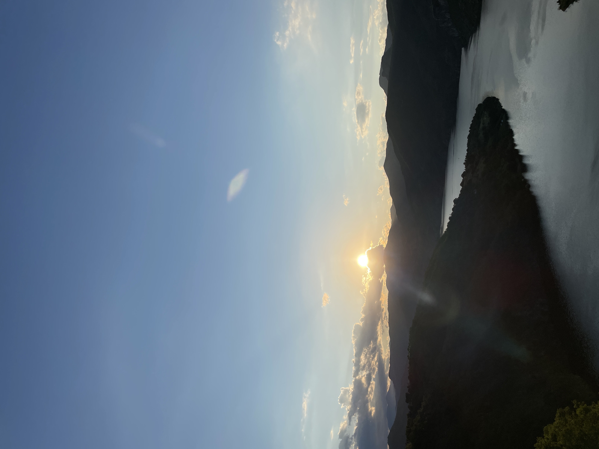 The image depicts a scenic view of a river winding through a valley surrounded by silhouetted mountains. The sun is setting behind the mountains, casting a warm glow on the clouds and the water. The sky is a gradient of blue, with scattered clouds. The foreground shows dark, tree-covered hills. This location is identified as the Borovitsa dam in Borovitsa, Bulgaria. There are no people or specific man-made objects visible in the scene, other than the implied presence of the dam structure which is not directly visible in this perspective. The environment is natural and vast, characterized by the interplay of light and shadow on the landscape.