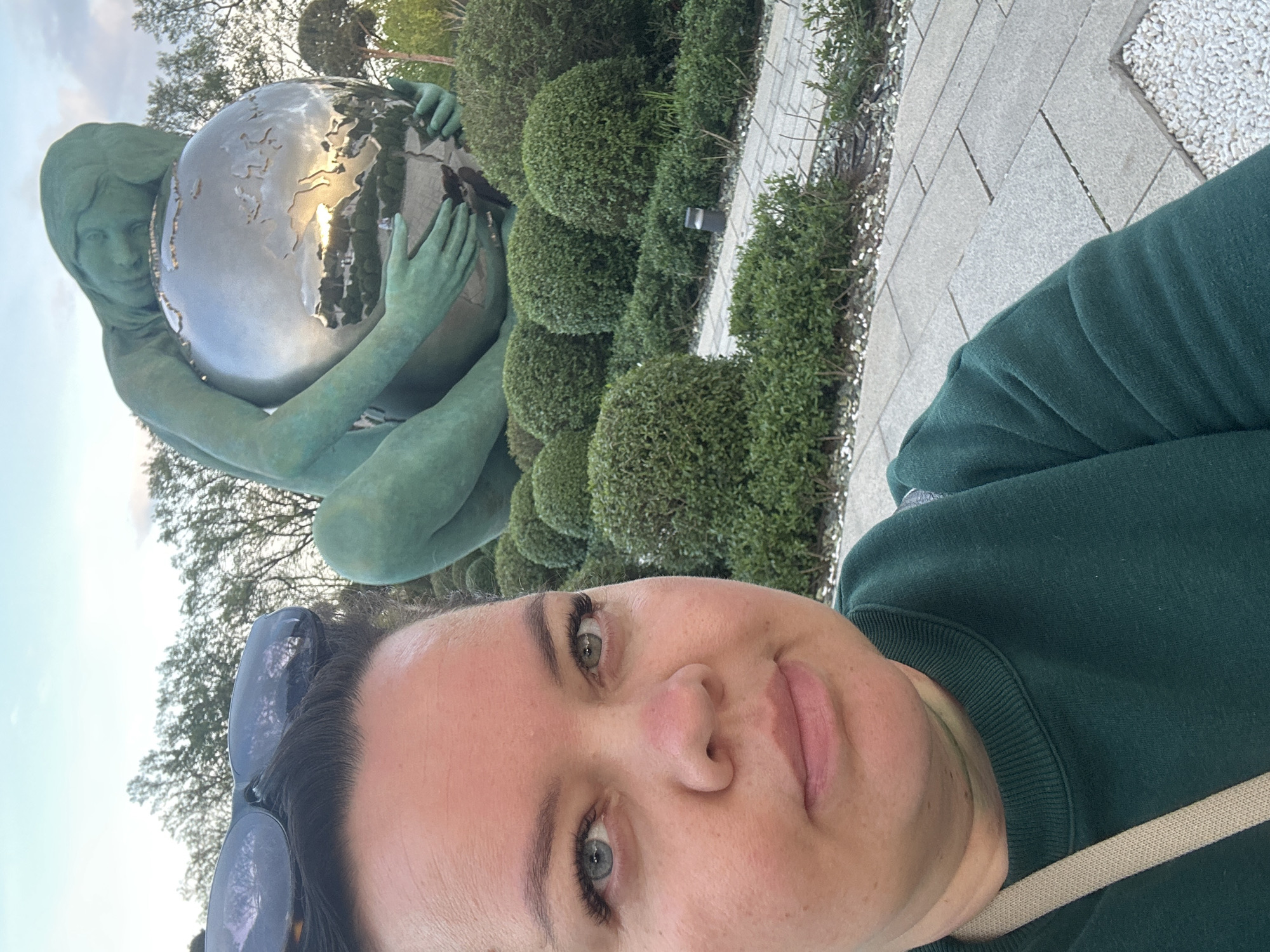 A woman is taking a selfie in front of the "World in the Hands of a Girl" sculpture, located in the Solnechny Ostrov Park in Krasnodar, Russia. The sculpture depicts a green-colored woman holding a silver globe. Behind the sculpture are neatly trimmed, round green bushes. A paved walkway is visible on the right side of the image. The woman in the foreground is wearing a dark green sweater and has sunglasses pushed up on her forehead.