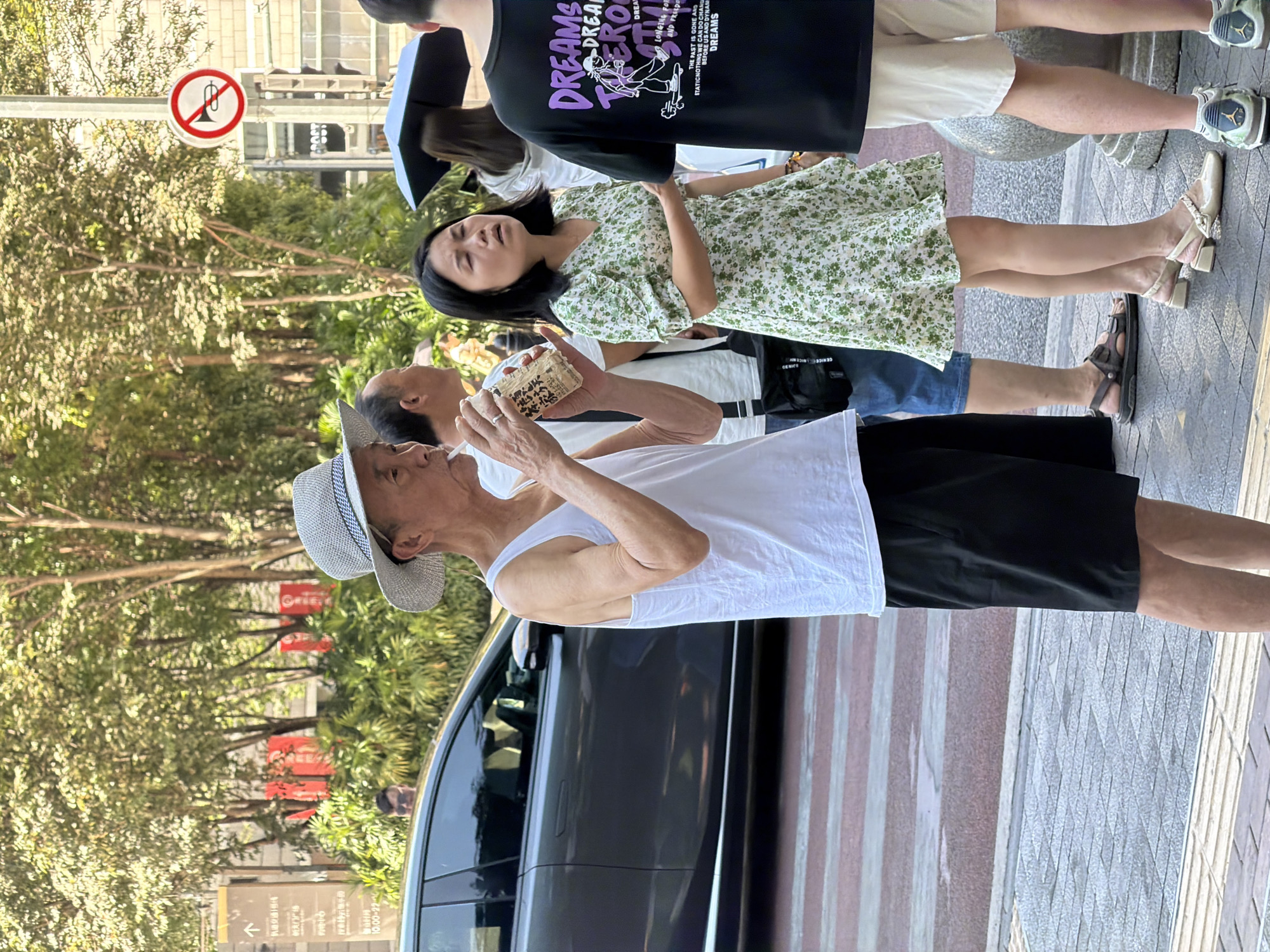 In Yuzhong District, China, an elderly man wearing a white tank top and black shorts stands on a sidewalk, holding a beverage and a straw to his mouth. He is wearing a straw hat and appears to be looking to his right. To his right, a woman in a green and white floral dress stands with her eyes closed, her mouth slightly open. Behind her, a person wearing a black t-shirt with purple text is visible. In the background, trees line the street, and a dark gray car is parked. A traffic sign with a red circle and a line through a horn symbol is posted on a pole. A crosswalk is present in the foreground.