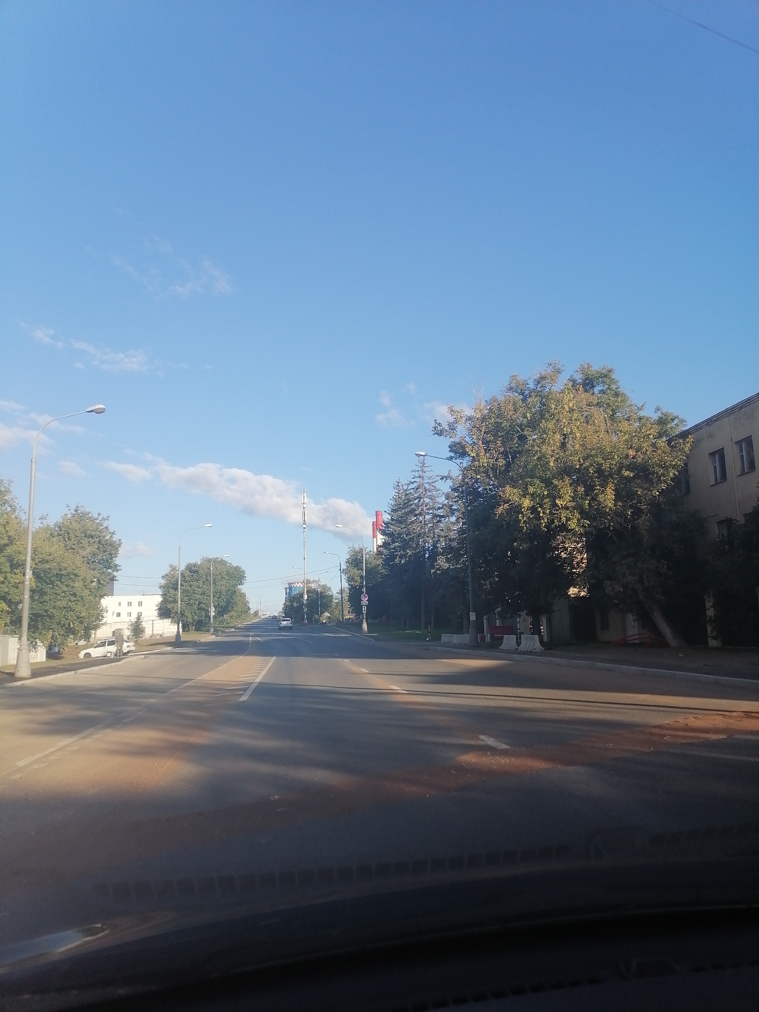 The image depicts a street scene in an urban environment, likely a residential or industrial area, under a clear blue sky with scattered clouds. The perspective is from inside a vehicle, looking forward.
In the foreground, the dashboard of a car is visible, along with the windshield. The road ahead is paved and appears to be a multi-lane street, possibly a highway or a main thoroughfare. Lane markings are visible, and there are some irregular reddish-brown patches on the road surface, possibly from spillage or staining.
On the left side of the road, there are several trees and a lamppost. Further down the street on the left, a white building with multiple windows is visible, and a few cars are parked. There's also a person standing near a white car.
On the right side of the road, there are more trees and a large building with windows. A tall communication tower or antenna structure is visible in the distance, with a flag on top. Red and white barriers are present along the roadside in some sections.
Several vehicles are seen driving on the road in the distance. The overall lighting suggests it is daytime, possibly morning or late afternoon, given the angle of the shadows cast by the trees. The environment appears to be orderly, with well-maintained infrastructure. The presence of trees suggests a developed urban area with green spaces.