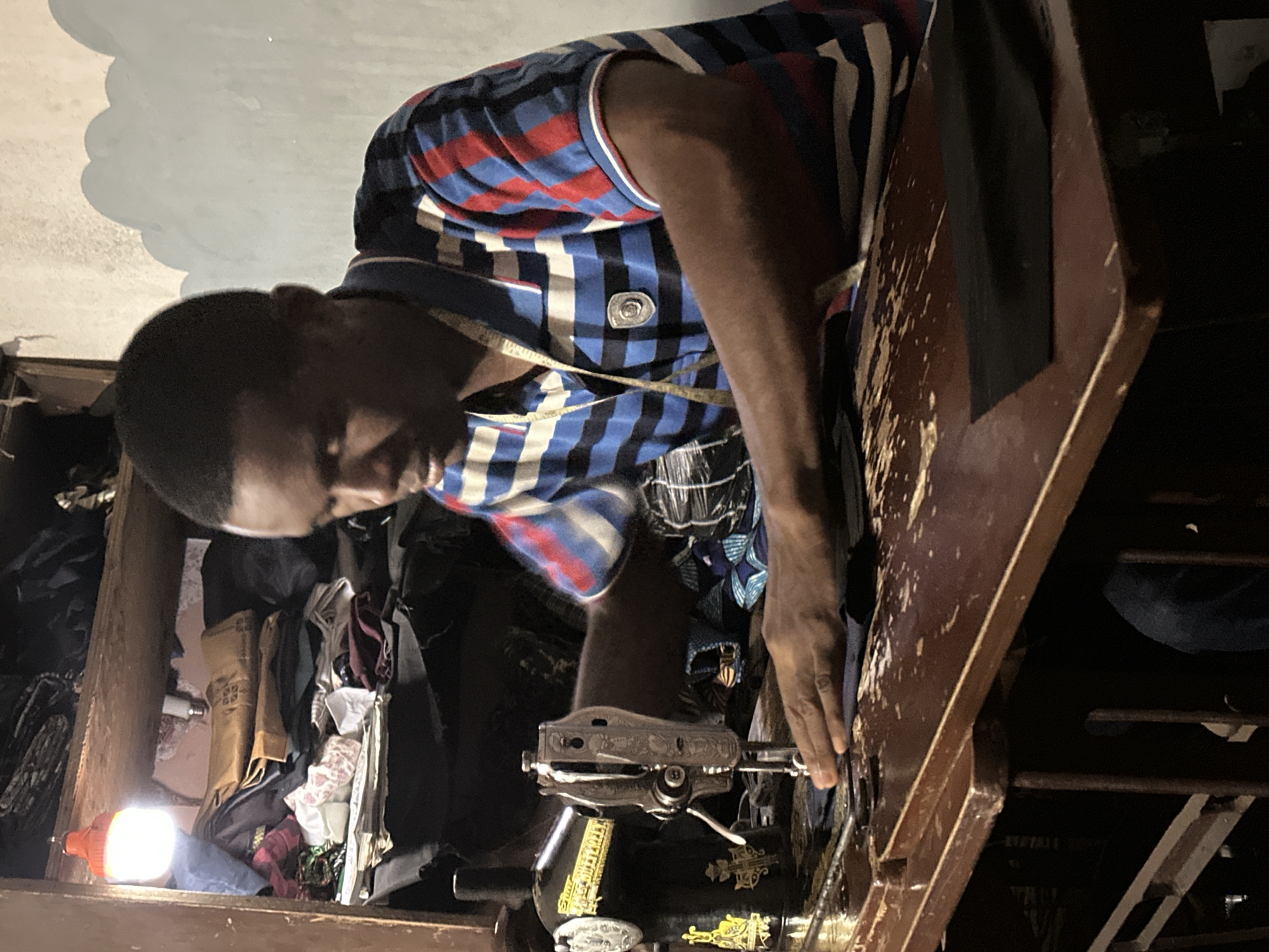 A man is pictured indoors operating a sewing machine. The man has short black hair and wears a striped, short-sleeved shirt with a measuring tape around his neck. He is focused on the sewing machine, his hand positioned on the fabric feed. The sewing machine is old and black, decorated with gold-colored designs. The table surrounding the sewing machine is made of wood and has visible signs of wear. The background is cluttered, featuring stored items and fabrics. A light source is present in the upper left corner. The location is unknown.