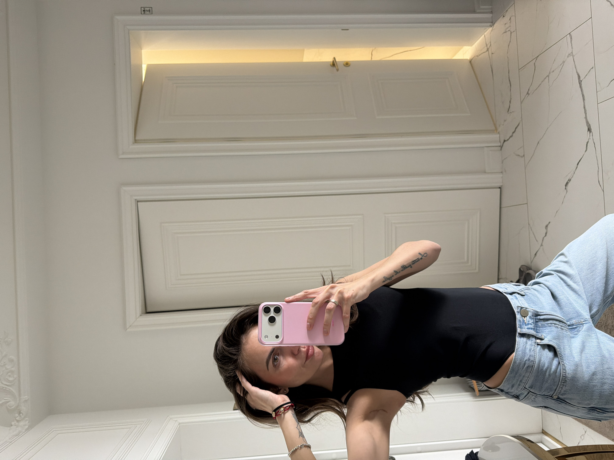A young woman captures a mirror selfie, angled gracefully as she holds a light pink smartphone with a triple-lens camera towards her reflection. She gazes directly at the phone's lens with a composed expression. Her hair is dark brown, and she is dressed in a black, form-fitting top and light-wash blue jeans. On her left hand, she wears a silver ring, and her left wrist is adorned with red and black string bracelets and silver beaded bracelets. Visible on her right arm is a tattoo reading "all things are possible to him that believeth."
The indoor setting features a clean, bright aesthetic, possibly a bathroom or a well-lit modern space. The background primarily consists of white walls and white doors with classic paneling. Two doors are prominent, one seemingly above the other, with the upper door slightly ajar. A warm, yellow light strip is visible above the upper door, illuminating the space. The right side of the image reveals a white surface with grey veining, characteristic of marble or a marble pattern, covering the wall and potentially the floor. Decorative white molding is visible on the lower left wall. A small, dark symbol resembling a building icon is etched into the top frame of the upper door. The scene conveys a sense of contemporary interior design. This location is identified as Krasnoyarsk, Russia.