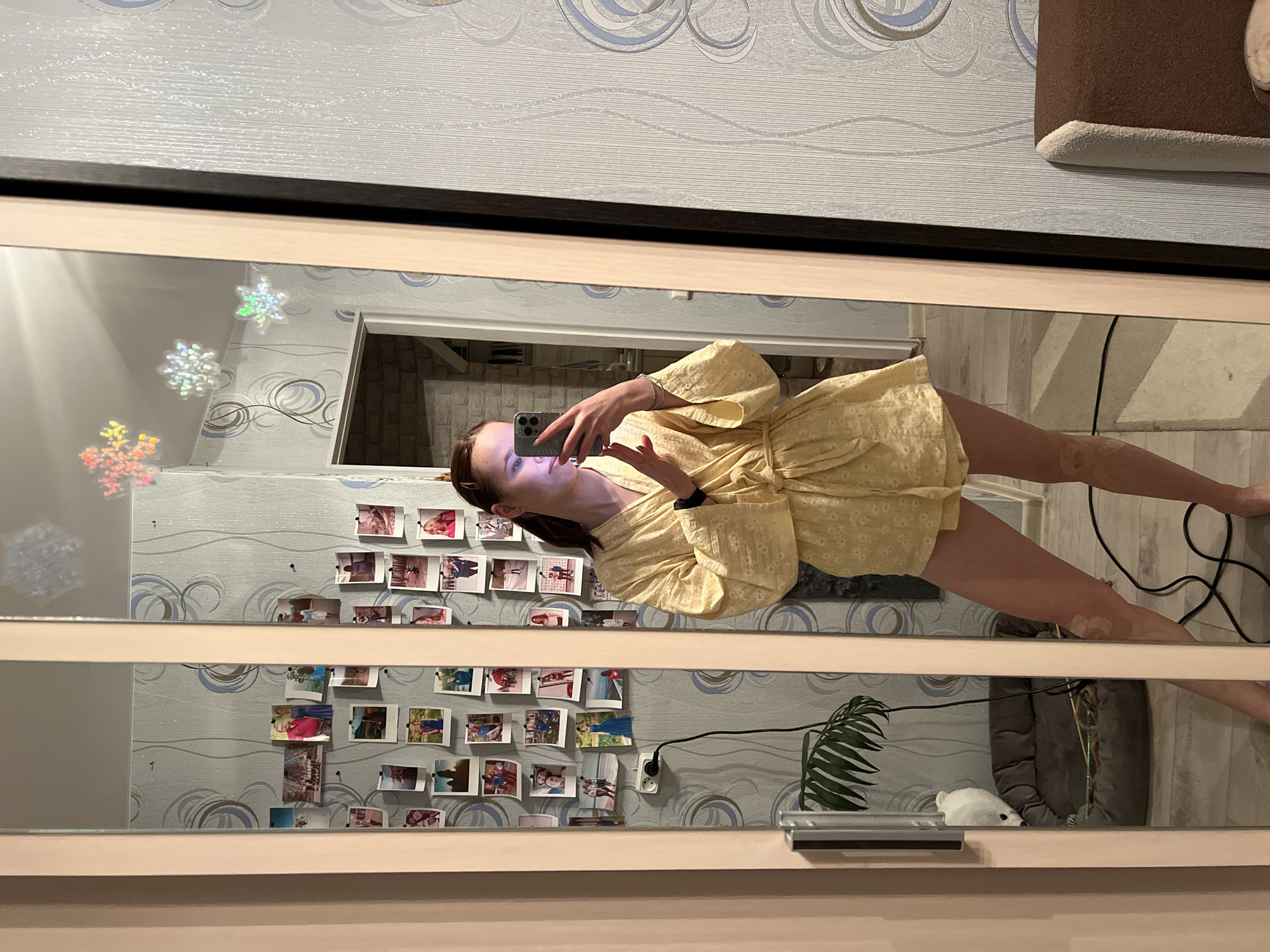A woman in a yellow dress is taking a photo of herself in a mirror. The mirror is mounted on a wall with decorative snowflakes and photos. A power outlet, a plant, and a white animal figurine are visible. The location is Chaikovsky, Russia.
