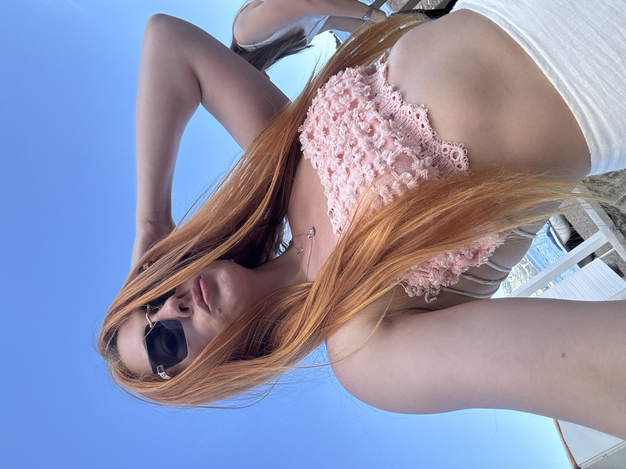 An inverted, close-up outdoor shot features a person with long, flowing reddish-blonde hair, likely a woman, against a clear blue sky. The perspective is from below, looking upwards. The woman is wearing dark sunglasses that obscure her eyes, and her lips are slightly parted. She has a small, silver necklace with a delicate pendant around her neck. Her top is a distinctive pink, possibly crochet-style, with numerous small, textured flower-like details. White straps are visible across her back.
In the upper background, partially obscured, is another person or part of a person wearing a white top. Elements of what appear to be light-colored outdoor furniture or structures are also visible in the background, suggesting an open-air setting. The bright, clear blue sky indicates a sunny day, likely midday or afternoon. The overall atmosphere suggests a leisurely moment in a warm climate. Given the context of Budva, Montenegro, the setting is likely a coastal or resort area, perhaps a balcony, terrace, or beachside lounge. No visible text is present in the image.