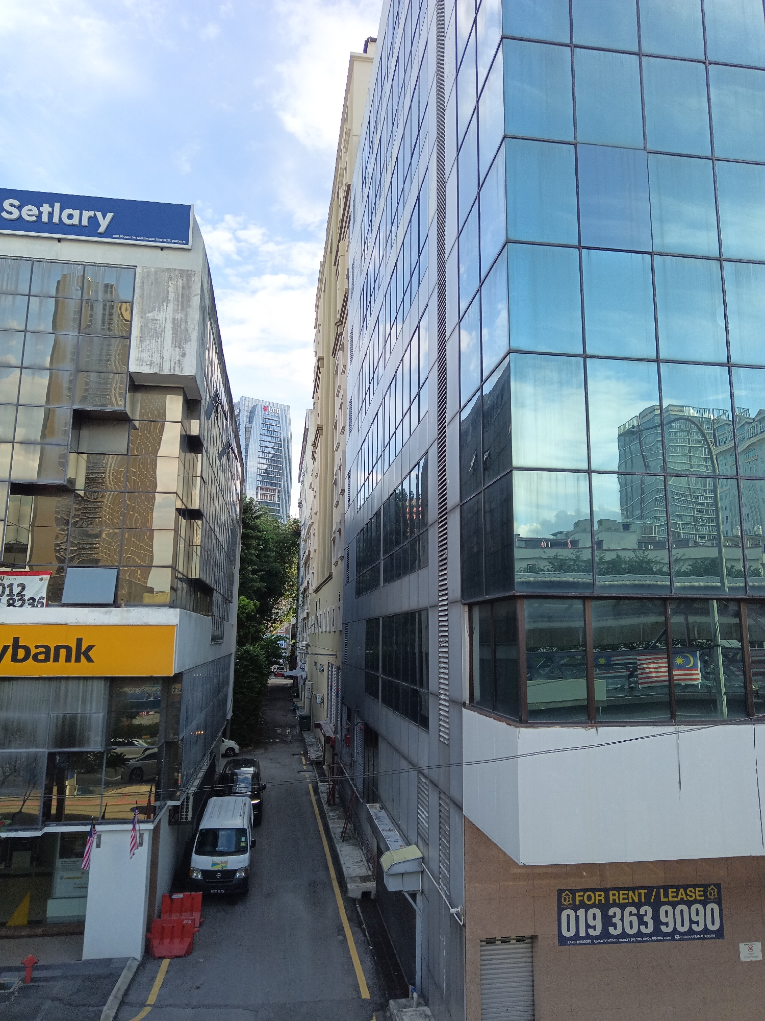 The image shows a street in Kuala Lumpur, Malaysia, with tall buildings on either side. The building on the left has a sign that reads "Setlary" in blue text, and below it, a yellow sign that says "aybank". The building on the right is a modern glass-fronted building. In the background, another tall building with a distinctive design is visible, possibly the Felda Tower. The street is narrow and appears to be an alleyway, with parked vehicles including a white van and a dark SUV. There are also some red traffic barriers and a fire hydrant visible. The sky is partly cloudy. The overall impression is of an urban environment with a mix of older and newer architecture.
