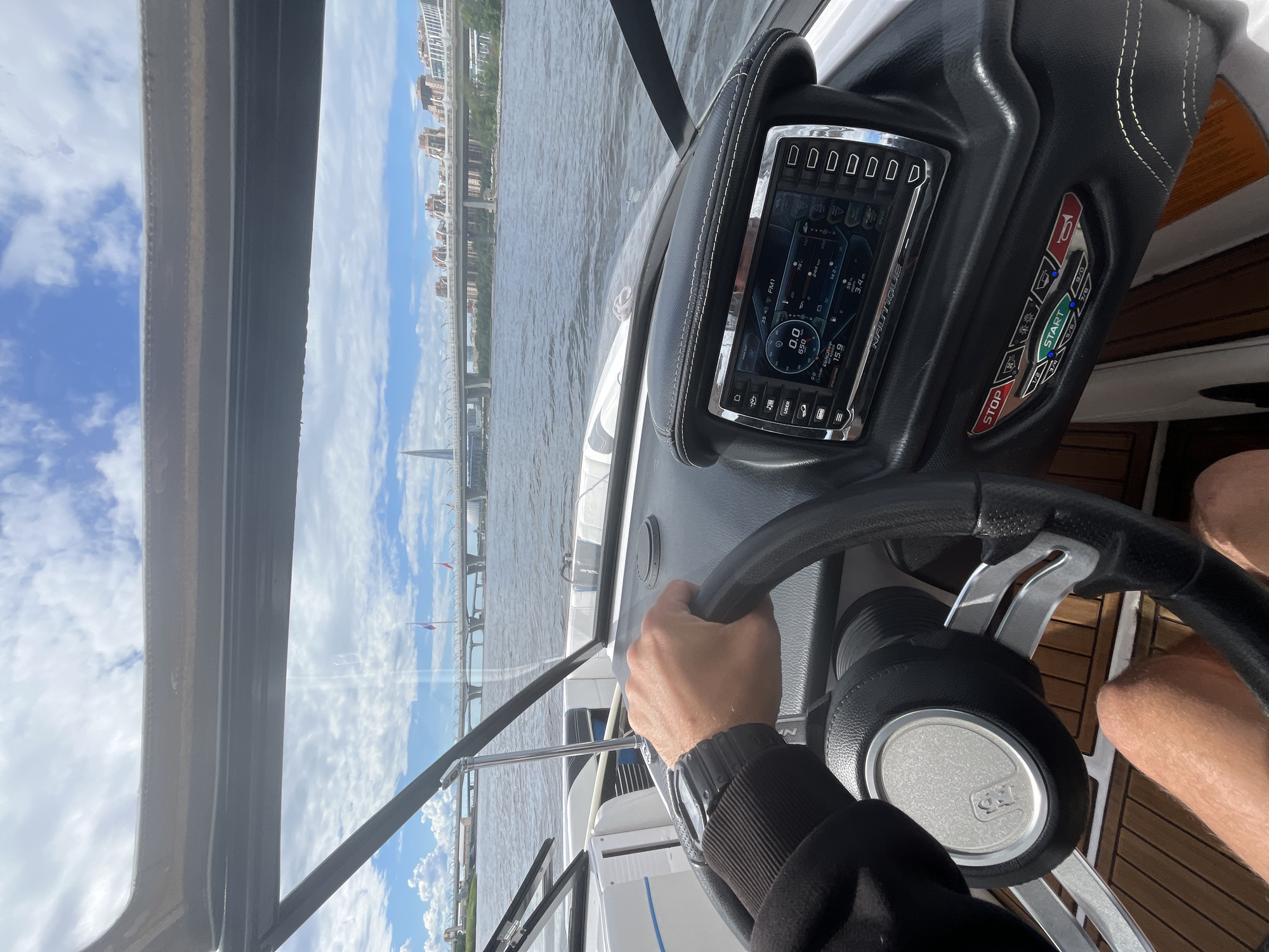 This media file shows a first-person perspective from the helm of a boat navigating a body of water. The view is directed forward and slightly to the right, revealing a modern boat console with a large digital display screen and various buttons. The screen shows navigation information, including speed (0.0 knots), RPM (650), and depth (3.4 m). It also displays icons and text related to boat functions and audio control, with "NAUTIQUE" visible on the screen bezel.
To the right of the screen are physical buttons labeled "STOP" and "START," with other indicators and options like horn and lights. The steering wheel, covered in black leather, is visible with a person's hand on it, indicating they are steering the boat. The person is wearing a watch on their left wrist and part of their legs are visible, suggesting they are seated.
The scene outside the boat is a waterway with a city skyline in the background. Buildings and bridges are visible along the shore. The water appears calm, and the sky is partly cloudy with blue patches, suggesting fair weather. The overall impression is a leisurely ride on a boat on a pleasant day, likely in an urban or suburban setting given the prominent city architecture. The time of day appears to be daytime, possibly late morning or afternoon due to the brightness. The presence of a modern boat and sophisticated dashboard suggests a recreational activity.