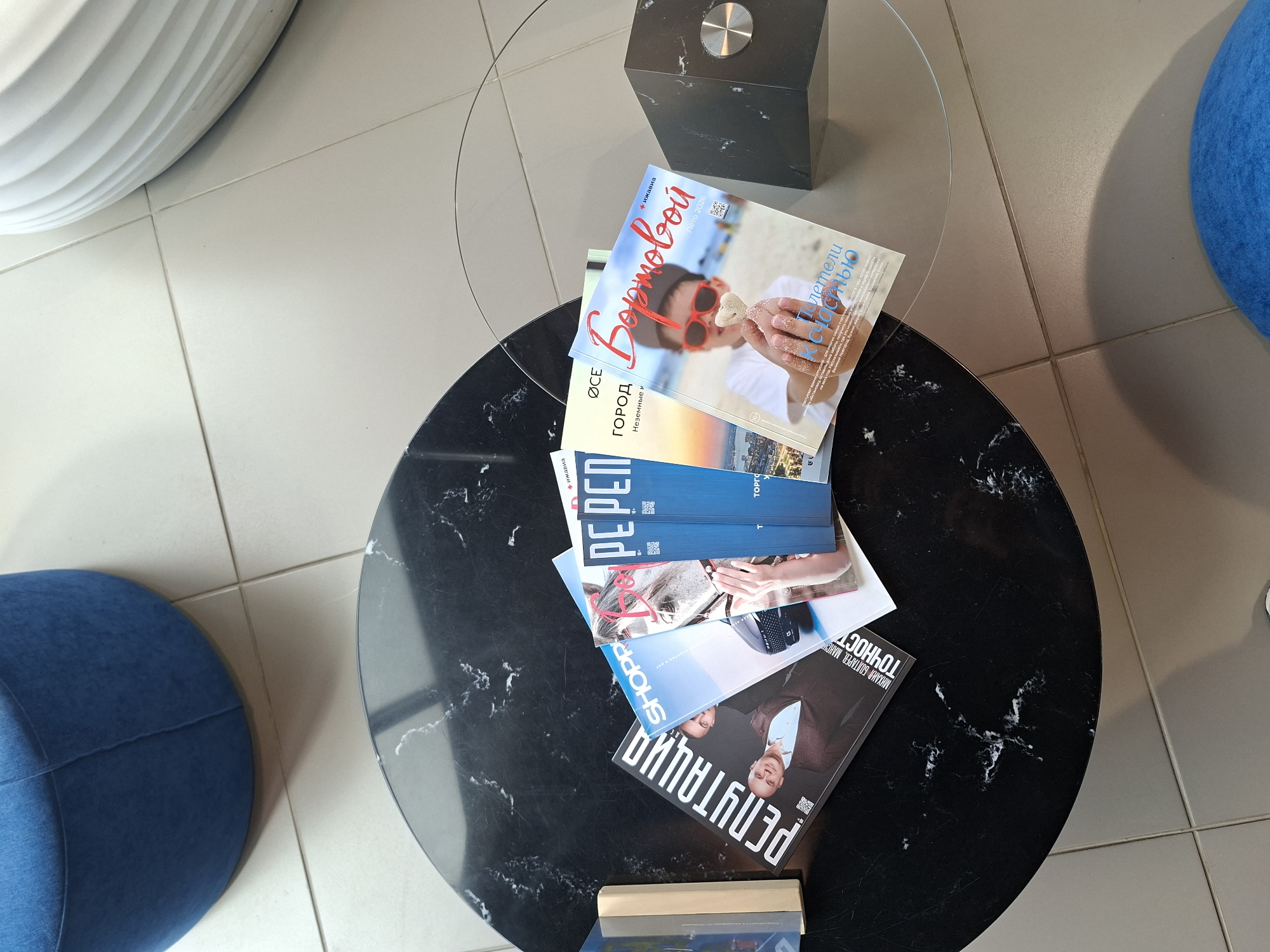 The image shows a collection of magazines on a black marble-patterned coffee table. The magazines are fanned out, with their covers visible. One magazine has the title "РЕПУТАЦИЯ" (Reputation) with a picture of a man on the cover. Another has "SHOPPR" in large letters. Several other magazines are partially visible, including one with a horse on the cover and one with the title "Вортовой" (Boarding) and an image of a child holding a cookie shaped like a heart. The magazines appear to be in Russian. The coffee table has a glass top. In the background, there is a blue upholstered ottoman and a ribbed white object, possibly another piece of furniture. The floor is tiled with light-colored tiles. The scene is indoors, likely in a waiting area or lounge. There is no specific indication of the location within the city of Izhevsk, Russia, other than the likely language of the magazines.