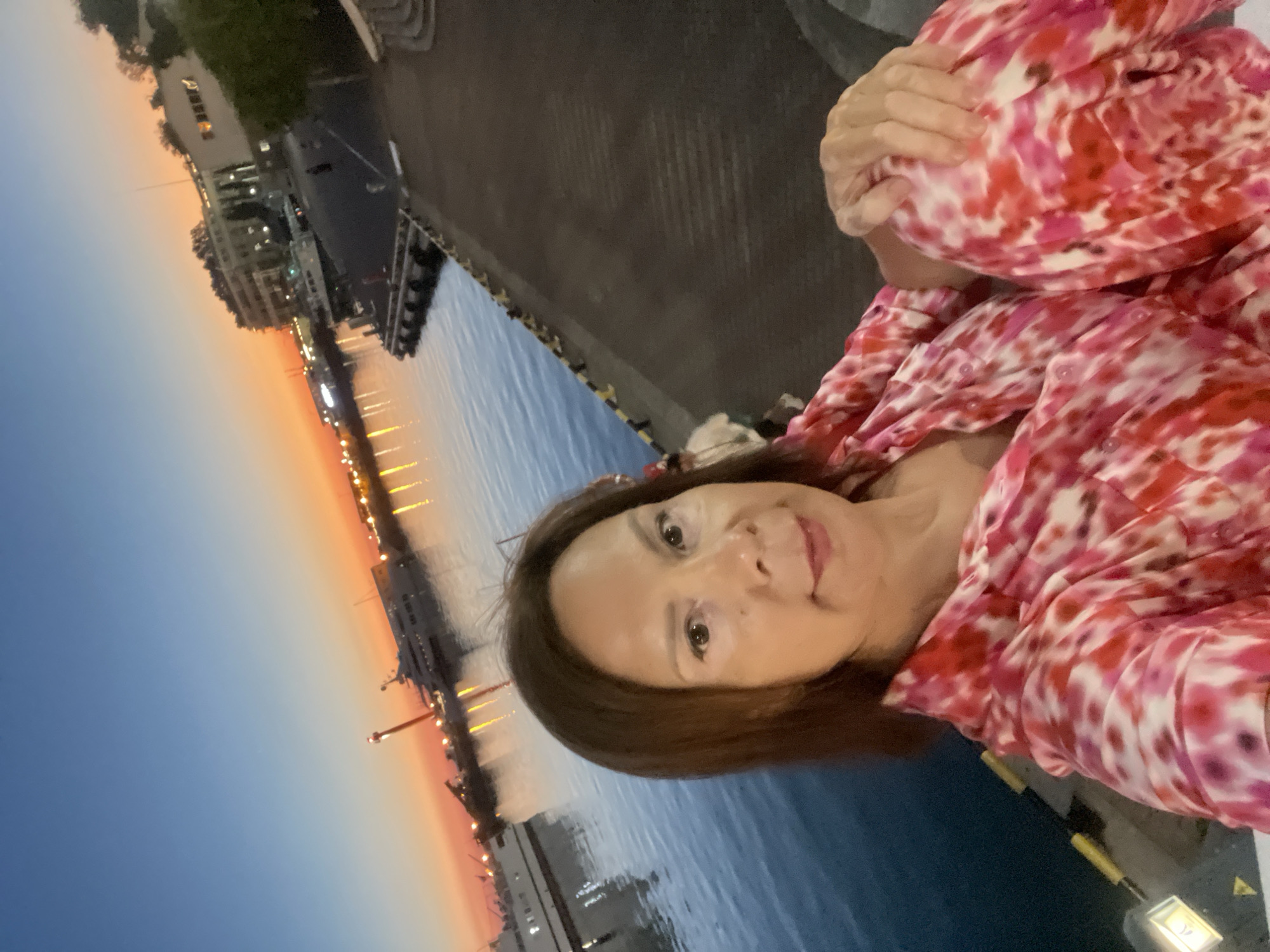 The image is a selfie taken outdoors at dusk, likely in a coastal city. The subject is a woman with shoulder-length brown hair, smiling at the camera. She is wearing a pink and white tie-dye patterned shirt. In the background, there is a body of water that appears to be the sea or a large bay, with several boats docked along the shore. Buildings with lights on are visible on the left side, suggesting an urban or port area. The sky displays a gradient of colors from deep blue at the top to orange and pink near the horizon, indicating sunset or twilight. The overall mood is peaceful and serene.