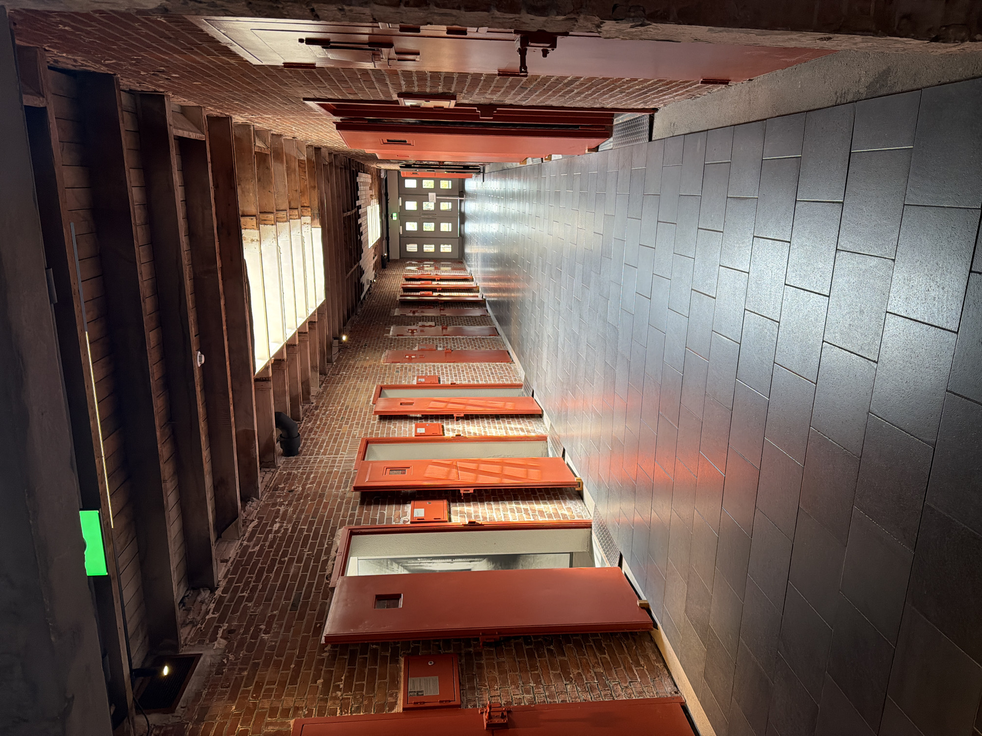 This is a photograph of a long, narrow corridor with an exposed wooden ceiling. The walls are made of red brick and are lined with identical red doors, each with a small square window. The floor is tiled with dark grey square tiles. Sunlight streams in from a skylight in the ceiling, casting reflections on the floor. A bright green illuminated sign is visible on the ceiling near the top of the image. The scene evokes the atmosphere of a prison or an old industrial building. The location is the former Bijlmer prison in The Hague, Netherlands, which has been converted into a cultural space. There are no people visible in the image.