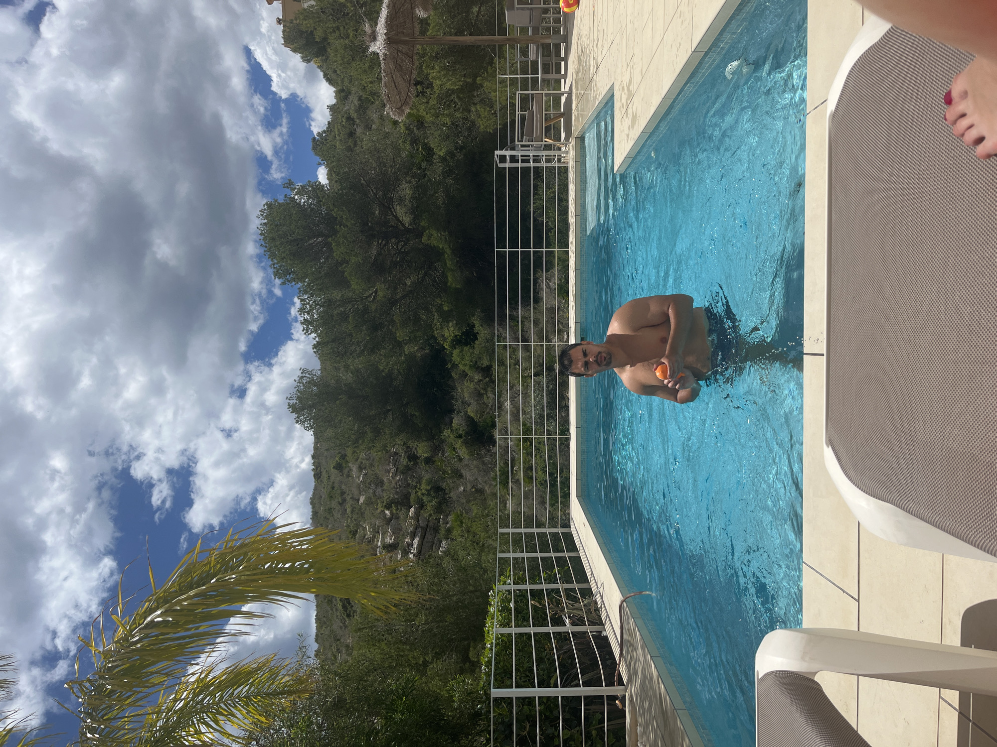 A man stands in a swimming pool, holding an orange ball. The pool is surrounded by white tiles and a white metal fence. Beyond the fence, lush green vegetation covers a hillside, with a partially visible rocky cliff in the background. The sky is blue with scattered white clouds. A palm tree is visible in the lower left corner. A sun lounger is positioned on the right side of the frame, partially cut off, with a person's foot visible on it. The scene suggests a private residence or vacation rental in a hilly, green area of el Poble Nou de Benitatxell / Benitachell, Spain, given its architectural style and vegetation.