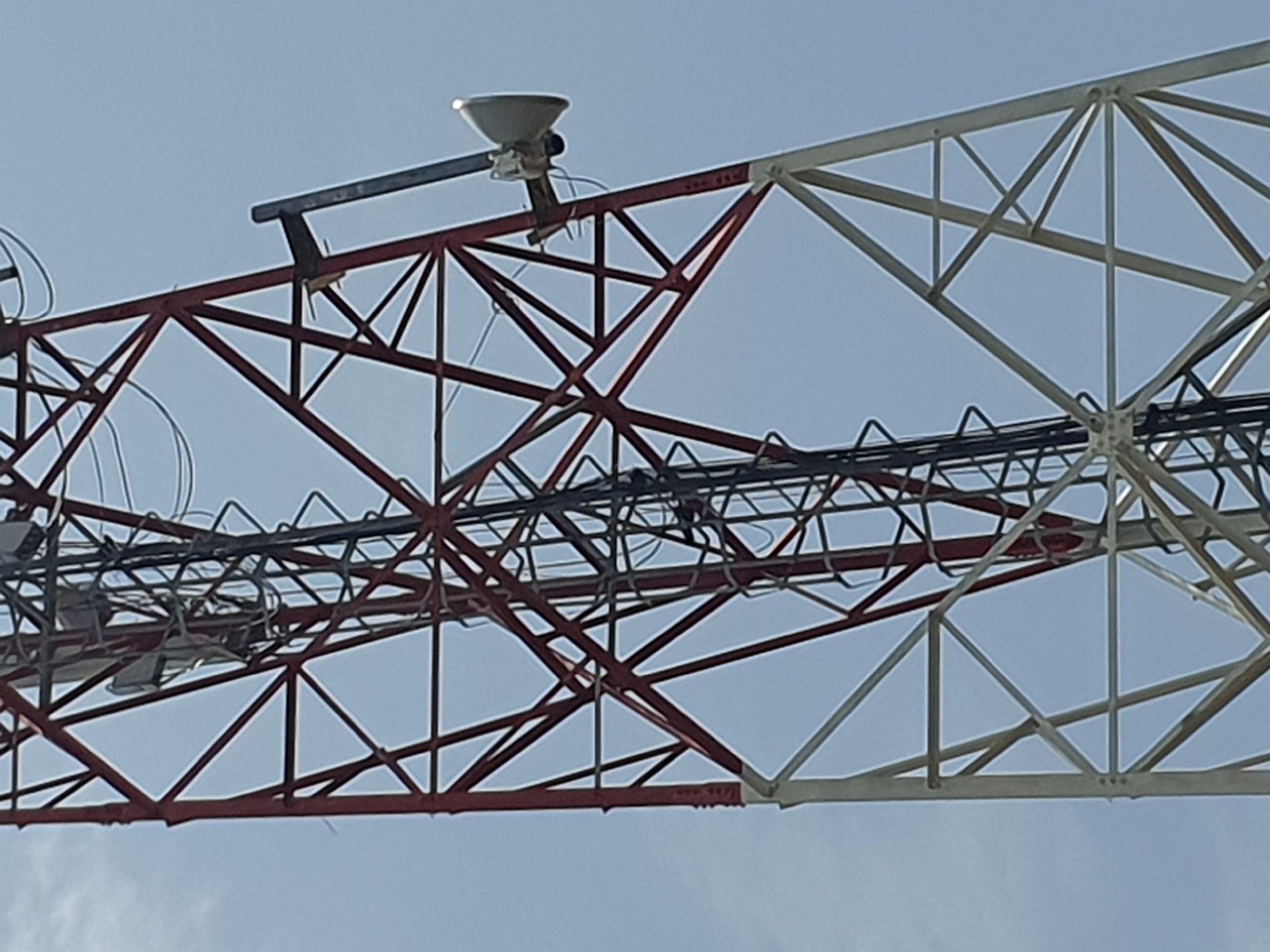 The image shows a close-up view of a red and white lattice tower, likely a telecommunications tower, against a pale blue sky. The tower is constructed from crisscrossing metal beams. A ladder runs vertically along the center of the tower. On the right side of the tower, a white parabolic antenna is mounted. The tower appears to be located in an open area, with no other structures or vegetation visible in the frame. The image does not provide any specific clues about the location being Gura Top, Nigeria, or any other details about the surrounding environment beyond the sky.