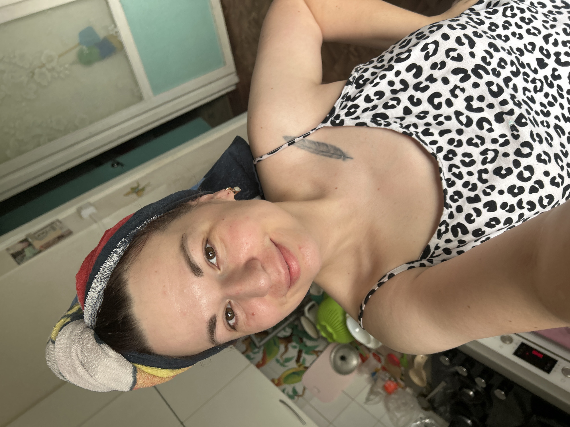 A young woman with brown hair tied up in a colorful towel is posing for a selfie in what appears to be a kitchen. She is wearing a white and black leopard print camisole. Her left arm is bent and resting on her hip, while her right arm is out of frame. A faint feather tattoo is visible on her upper chest. The background shows kitchen cabinets, a stove, and a window with a view of what looks like a room with blue and white walls and some hanging objects. The overall environment suggests a domestic setting. There is no indication of the specific location within the city of Fokino, Russia.