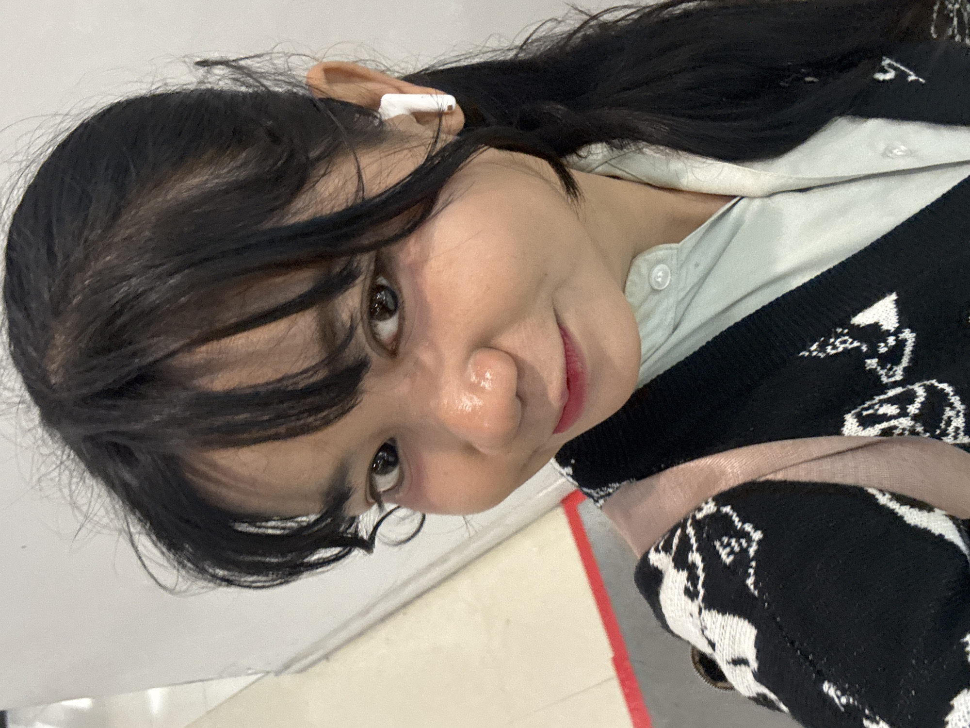 A young Indonesian woman of East Asian descent is captured in a close-up, selfie-style photograph. She has dark, shoulder-length hair with bangs, and her face is turned slightly to her left. She is wearing a light blue collared shirt beneath a black and white patterned cardigan. A light pink strap, likely from a backpack or purse, is visible over her right shoulder. A white wireless earbud is present in her right ear. The background is blurred and consists of a light-colored wall on the right and a lighter, off-white wall on the left, with a hint of a red line on the floor in the lower left corner. The lighting appears to be artificial, creating some shine on her face. The scene is likely indoors, possibly in a public space like an airport or mall, given the presence of the red line which could indicate a pathway or barrier. The overall impression is casual and everyday.