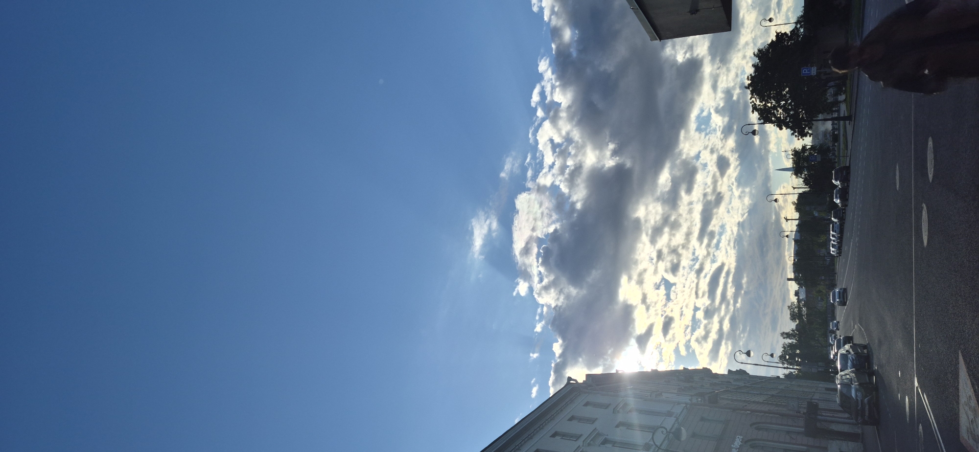 The image depicts a view of a city street in Saint Petersburg, Russia, likely during late afternoon or early evening, given the angle of the sun and cloud formations. The sun is partially obscured by dramatic, textured clouds, casting rays of light through them and onto the sky. On the left side of the frame, a large, light-colored building with multiple windows and architectural details dominates the foreground. The building appears to be a historical structure with a classical facade.
The street in front of the building is paved and shows lane markings. Several cars are parked along the side of the street and others are driving. The street is lined with ornate lampposts. In the middle ground, more buildings and trees are visible, suggesting a densely built urban environment. Further in the distance, a slender spire can be seen piercing the skyline, characteristic of Saint Petersburg's architecture.
A person, appearing to be an adult, is walking away from the camera on the right side of the street. They are wearing dark clothing and their form is partially blurred, suggesting movement. The overall impression is an urban scene bathed in dramatic, atmospheric light, showcasing the architecture and street life of Saint Petersburg. The specific location is likely a busy thoroughfare within the city center, given the presence of significant buildings, traffic, and pedestrian activity.
