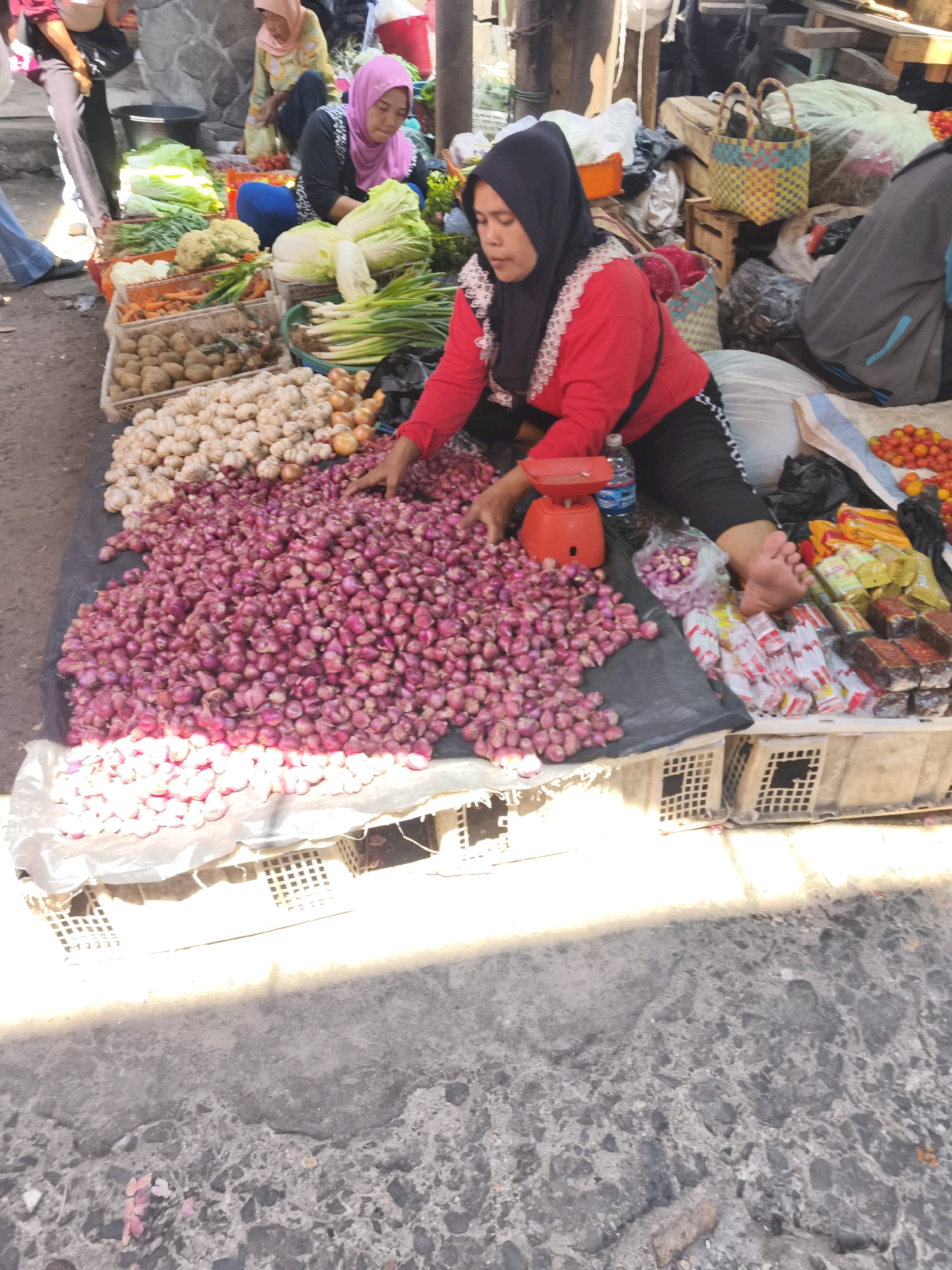 The image depicts a street market scene in Palembang, Indonesia, likely near a traditional market area like Pasar Gubah or Pasar 16 Ilir. In the foreground, a woman wearing a pink hijab and a red shirt is seated on the ground, surrounded by large piles of red onions. To her right, a red digital scale sits on a black cloth, suggesting she is weighing produce for sale. A bottle of water is also visible nearby.
Behind the woman, various fresh vegetables are displayed in crates. These include cabbages, carrots, potatoes, and garlic. Other individuals can be seen in the background, some of whom appear to be vendors or shoppers. The ground is paved with rough asphalt. The overall atmosphere suggests a busy, open-air marketplace with a focus on fresh produce.