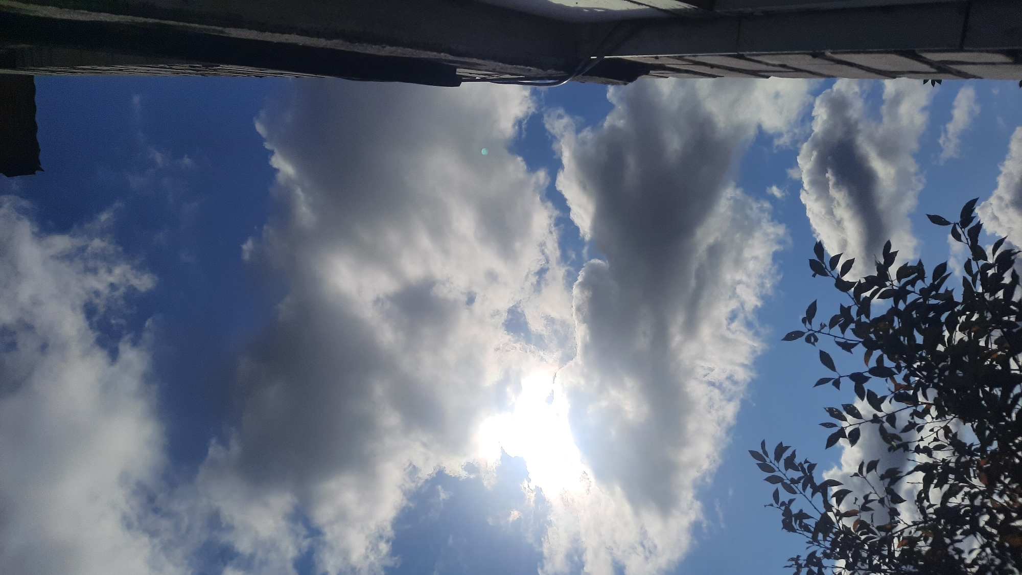 The image shows a view of the sky from a low angle, with a dark silhouette of tree branches in the foreground on the left. The sky is a deep blue, interspersed with a mix of bright white and dark grey clouds. The sun is partially obscured by a large, bright cloud formation, creating a lens flare effect and casting bright light. On the right side of the image, the corner of a building is visible, with a textured grey facade and an overhanging eave. The image appears to have been taken outdoors in an urban setting in Shkloŭ, Belarus. There are no people or animals visible in the image. The scene depicts a partly cloudy sky with sunlight breaking through, observed from ground level with part of a building and foliage framing the view.