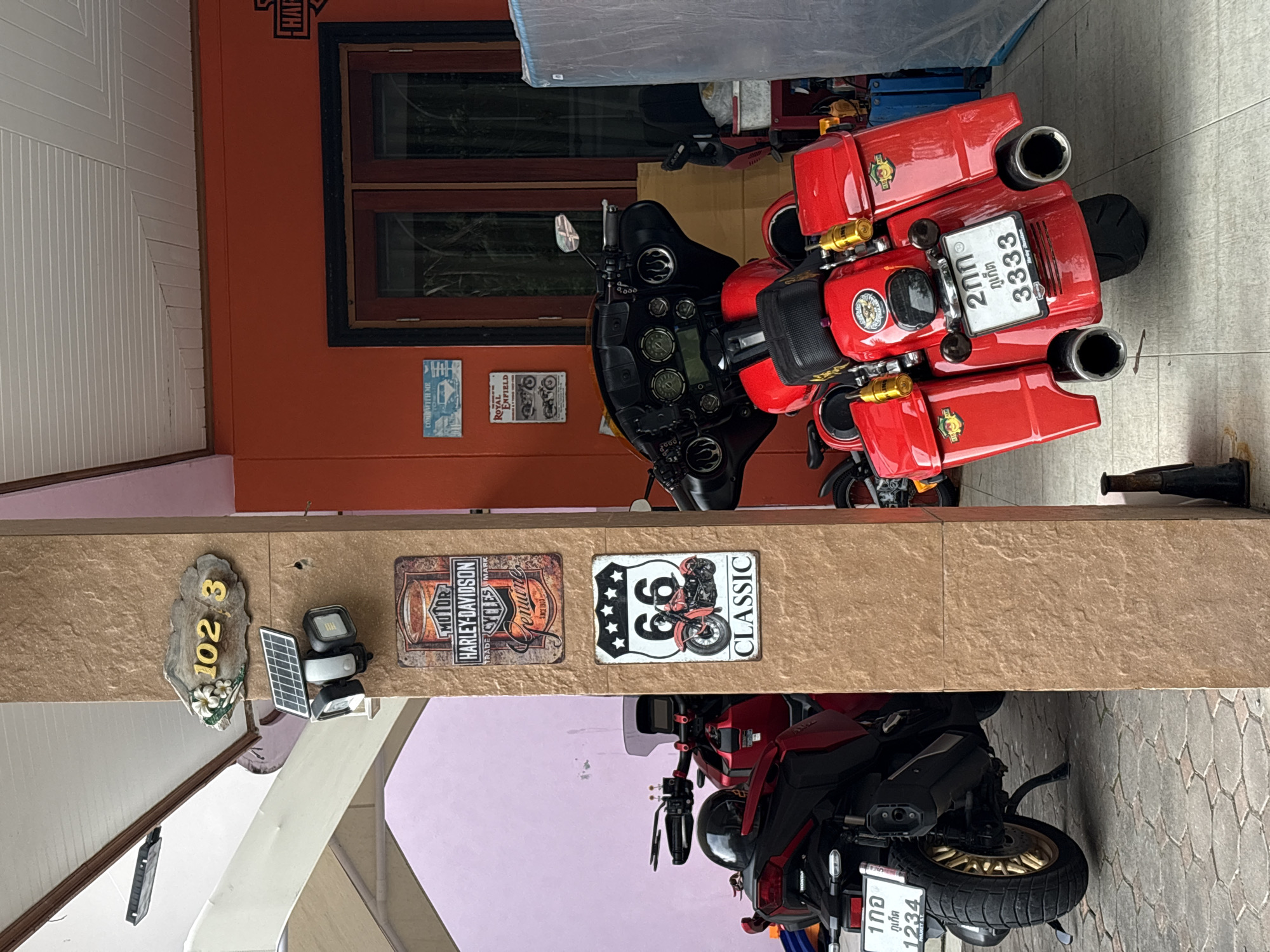 The image depicts an outdoor scene, likely a residential property, in Choeng Thale, Thailand. The foreground features two red motorcycles parked under a covered area. The motorcycle on the left is a scooter-style with a black seat and a gold-colored wheel, displaying a license plate with Thai characters and the number "1234". The motorcycle on the right is a larger cruiser-style bike, also red, with saddlebags and a license plate featuring Thai characters and the number "3333".
A pillar of the building, textured with a light brown stone pattern, stands prominently in the center of the image. Attached to this pillar are several decorative signs. The uppermost sign is a weathered-looking metal sign with "MOTOR HARLEY-DAVIDSON CYCLES GENUINE" printed on it. Below this is a classic Route 66 sign featuring the number "66" and a depiction of a motorcycle. Further up the pillar, a small solar-powered light fixture is mounted. Above the light, a decorative house number sign reads "102 3" with floral embellishments.
The background reveals parts of the building's exterior, painted in vibrant orange. A set of wooden double doors with dark frames is visible. To the left of the pillar, a section of a building with a light purple wall is seen. The ground is paved with hexagonal stones. Additional signs are visible on the orange wall, including one with a vintage car and the text "COME WITH ME" and another advertising "ROYAL ENFIELD". The overall impression is a well-decorated outdoor space with a clear emphasis on motorcycle culture.