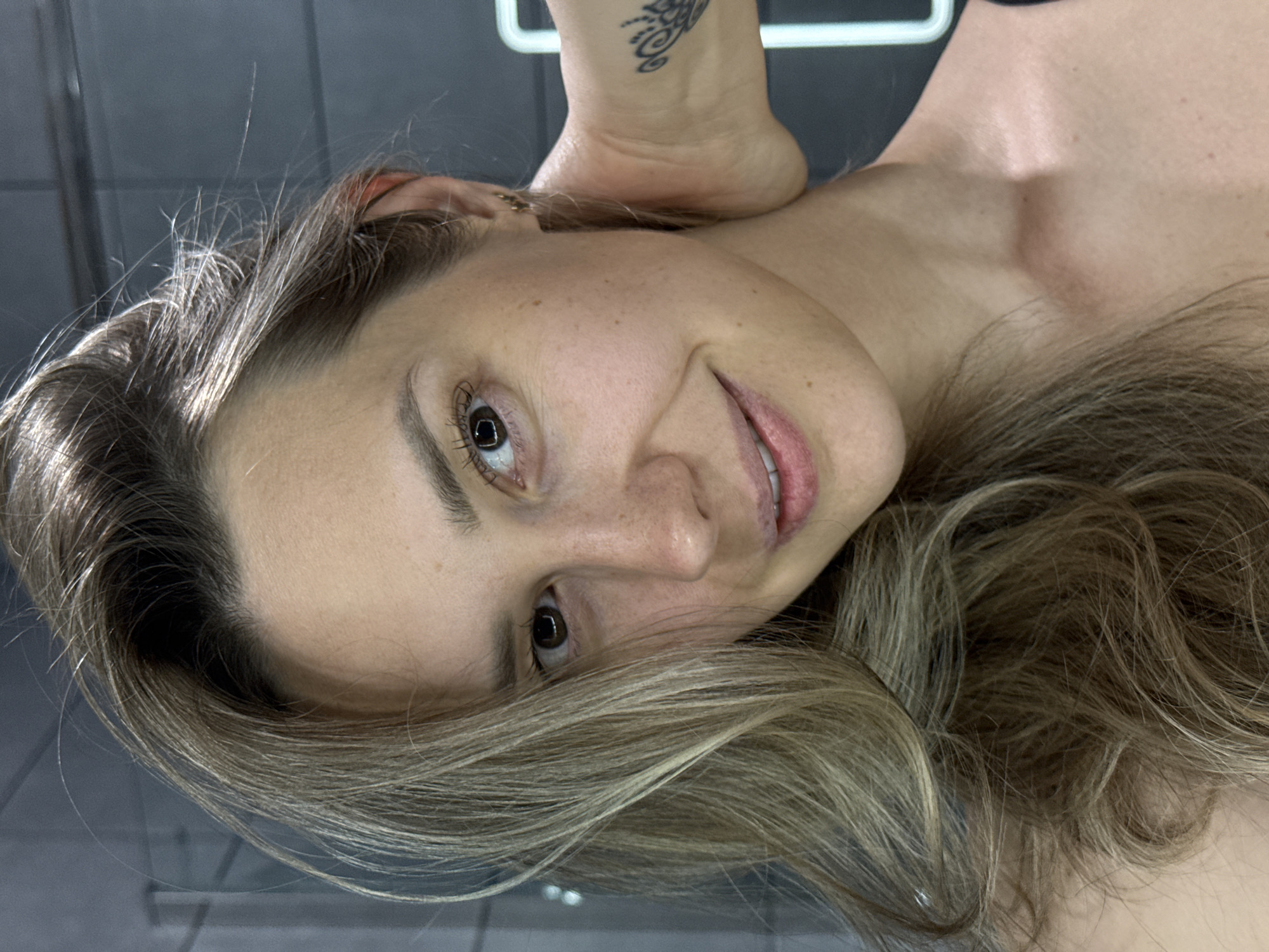 This close-up portrait, oriented sideways, features a woman with light skin, dark brown eyes, and a gentle, natural smile. Her long, wavy hair, a blend of light brown and blonde hues, frames her face and flows around her head, with some fine flyaway hairs visible near her hairline. Visible freckles dot her nose and cheeks.
Her gaze is directed towards the camera, conveying a relaxed and pleasant expression. Above her head, a part of her arm or wrist is visible, adorned with an intricate dark-patterned tattoo. She appears to be bare-shouldered or wearing clothing that reveals her shoulders and collarbone.
The background is dark and out of focus, suggesting an indoor setting. A white, linear object, possibly a handrail or part of a fixture, is partially visible in the upper background. The lighting highlights her hair and facial features, indicating an artificial light source. There is no visible text in the image. The provided context places this scene in Chaikovsky, Russia.