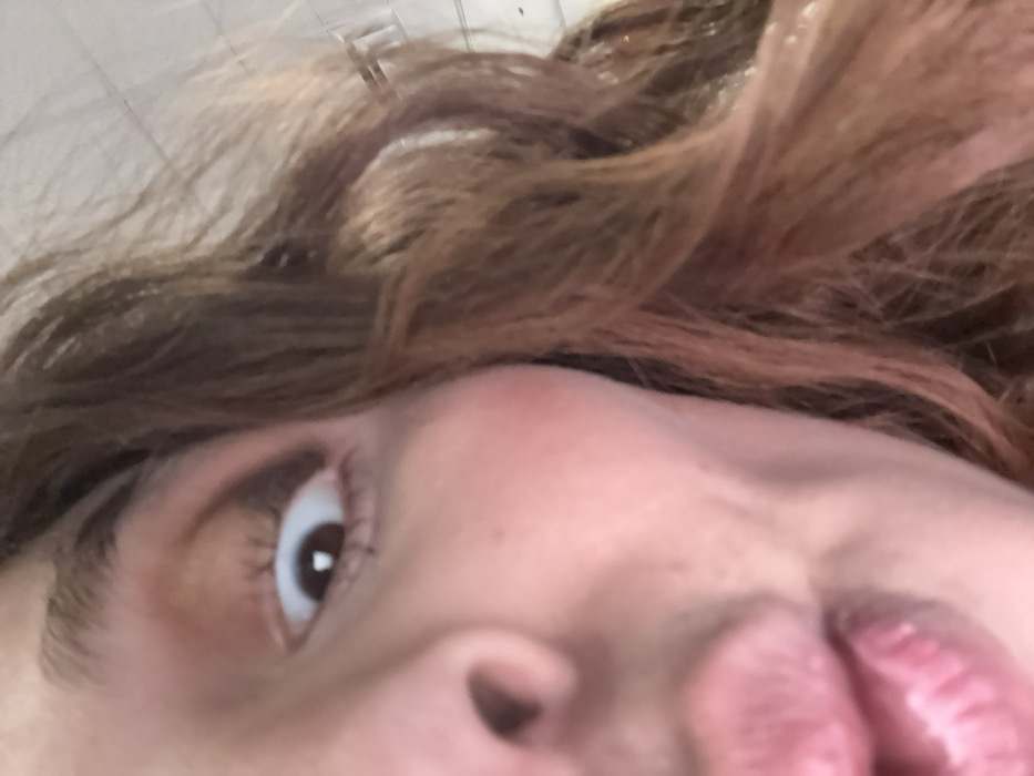 An extreme close-up focuses on a person's face from a low angle. The view includes the right eye, part of the nose, and mouth. The person has fair skin, brown hair, and dark eyelashes. The lips are pink and slightly parted. The eye looks upwards, showing the iris and a small glint of light reflecting off the pupil. The perspective and proximity create a distorted view of the facial features.