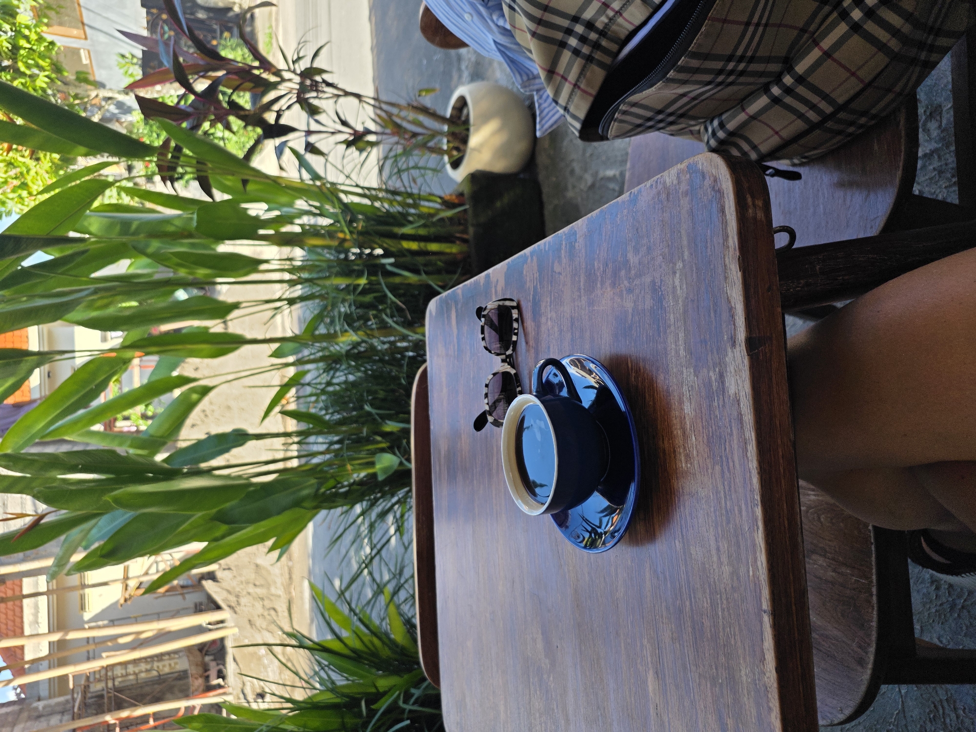 The image depicts a table setting at an outdoor café in North Kuta, Indonesia. A dark blue coffee cup and saucer sit on the wooden table alongside a pair of sunglasses. Lush green plants are visible in the background, and the architecture of nearby buildings is partially visible. In the background, a portion of a person with a plaid backpack and a planter are visible. A second person is seen sitting at the table, with their right thigh visible in the image. The scene suggests a casual, relaxed atmosphere.
