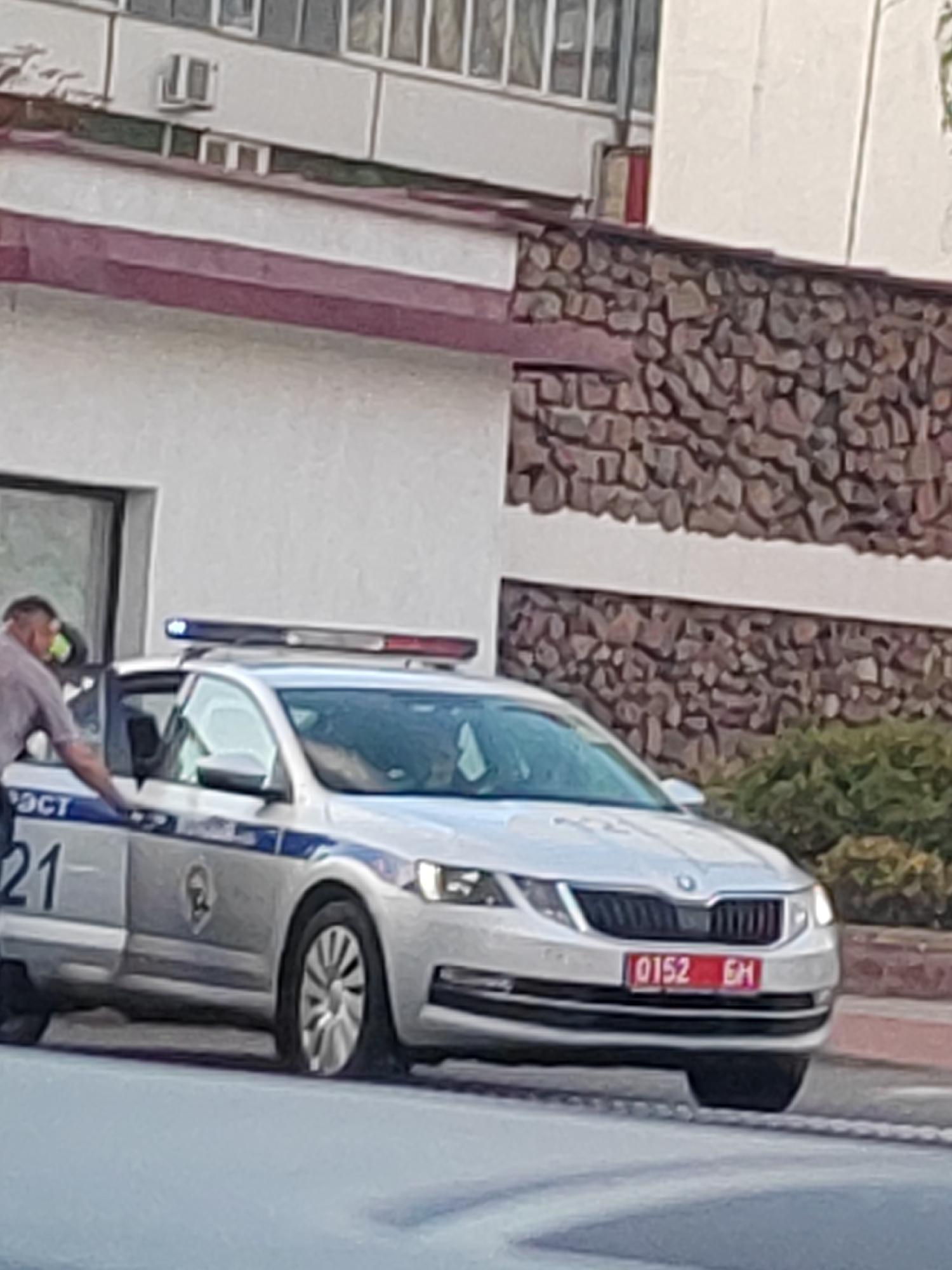 A silver Skoda Octavia police car with flashing blue and red lights is parked on a street. The car has a blue stripe along the side with Russian text and the number "21". The license plate reads "0152 EH". A man in a grey shirt is standing next to the driver's side door. In the background, there is a building with stone and white siding. Green bushes are visible behind the car.