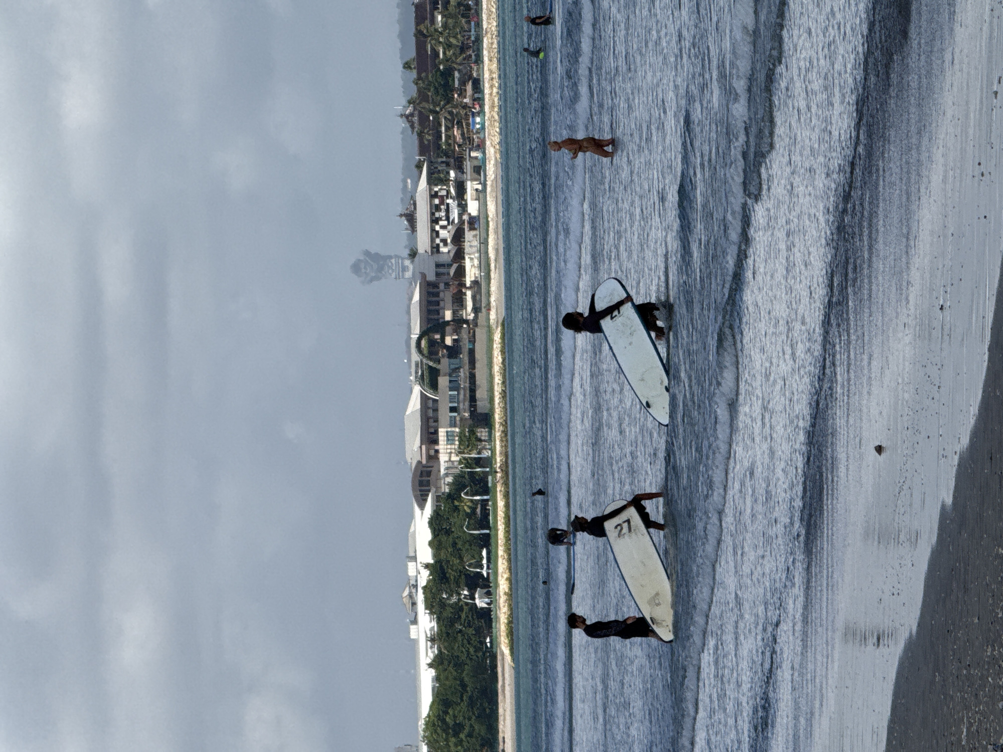 The image depicts a beach scene in Kuta, Indonesia. The photograph is oriented vertically and shows several people in the ocean, some with surfboards. The waves are visible and there's a sandy beach in the foreground. In the background, there's a line of buildings and vegetation characteristic of a populated coastal area, likely the urban part of Kuta. The sky is overcast, indicating possibly cloudy weather. Two surfers are visible with surfboards while one person is swimming or standing in the waves. The number "27" is visible on one of the surfboards.