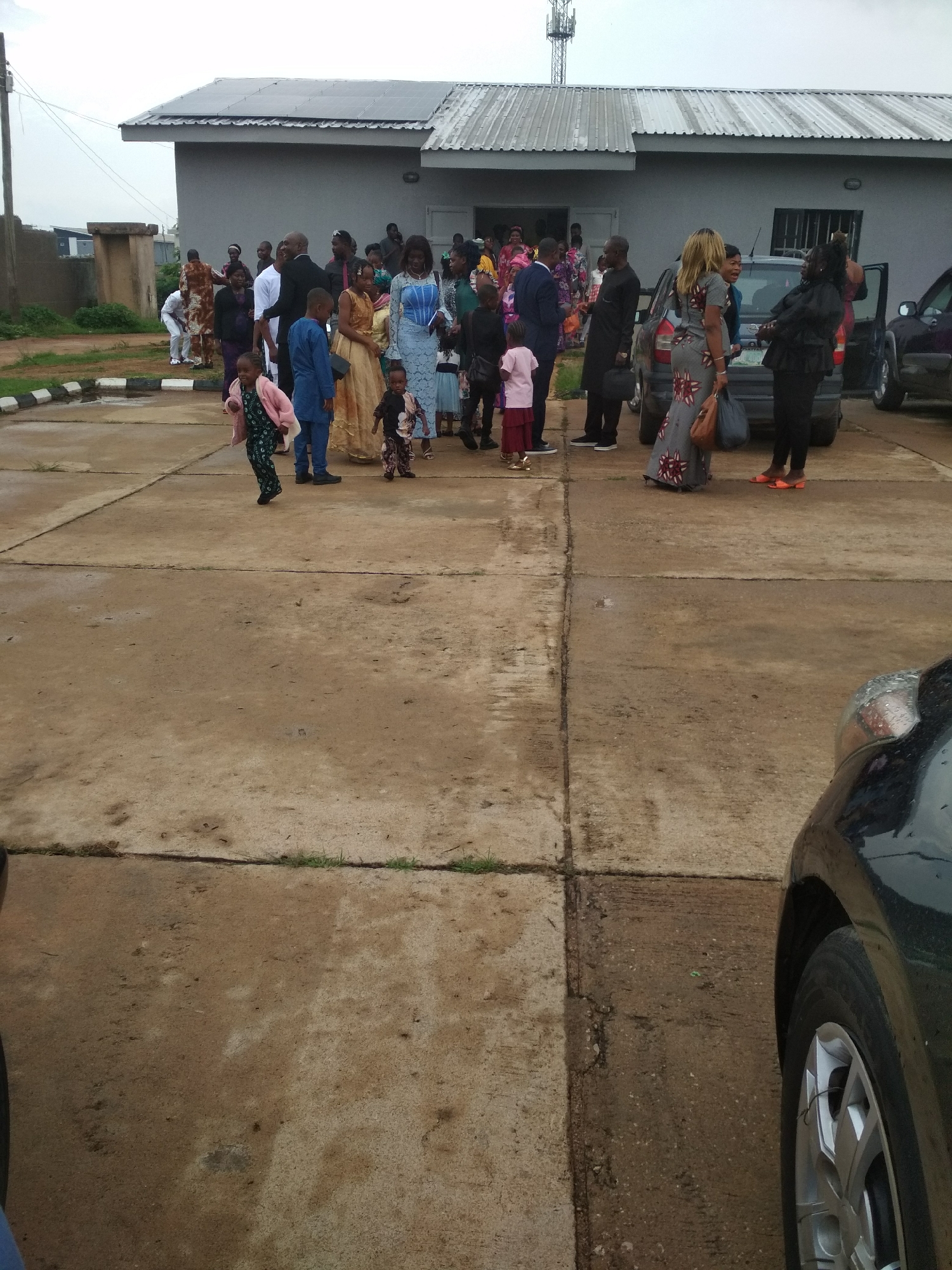 The image shows a group of people standing outside a light gray building with a metal roof and a telecommunications tower in the background, likely in the city of Jos, Nigeria. The group includes men, women, and children of various ages, some wearing formal attire. Several cars are parked nearby. The ground is concrete with visible seams and some patches of dirt and grass. The weather appears overcast. Some people are interacting and conversing, while others seem to be arriving or departing.