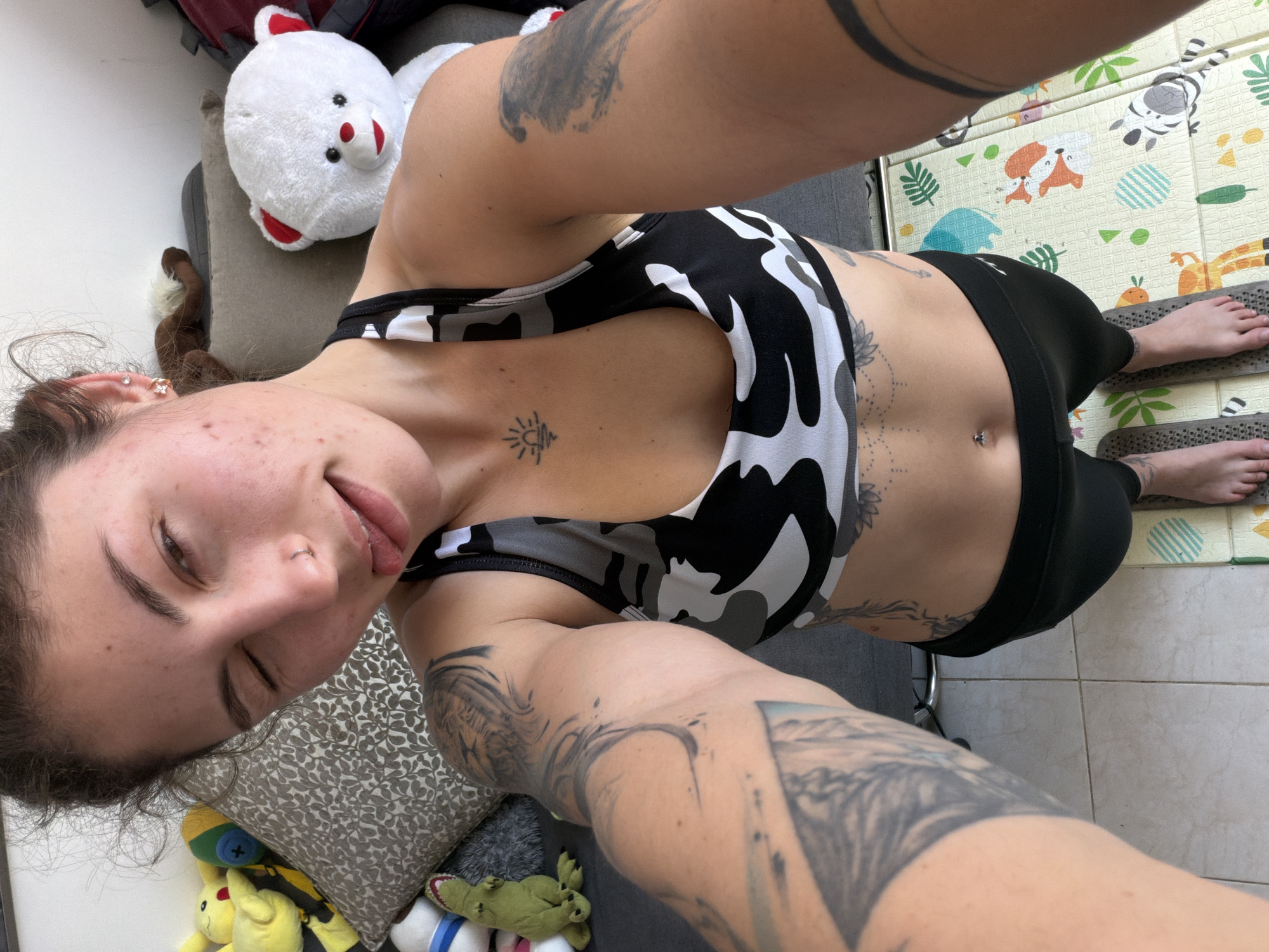 The image is a selfie taken indoors, possibly in an apartment in Kiryat Bialik, Israel. The woman is wearing a patterned sports bra and black leggings. She has multiple tattoos on her arms and stomach. She has a nose piercing and visible skin imperfections. She is surrounded by stuffed animals and a pillow on a grey couch. Her bare feet are visible on a play mat. The floor is tiled.